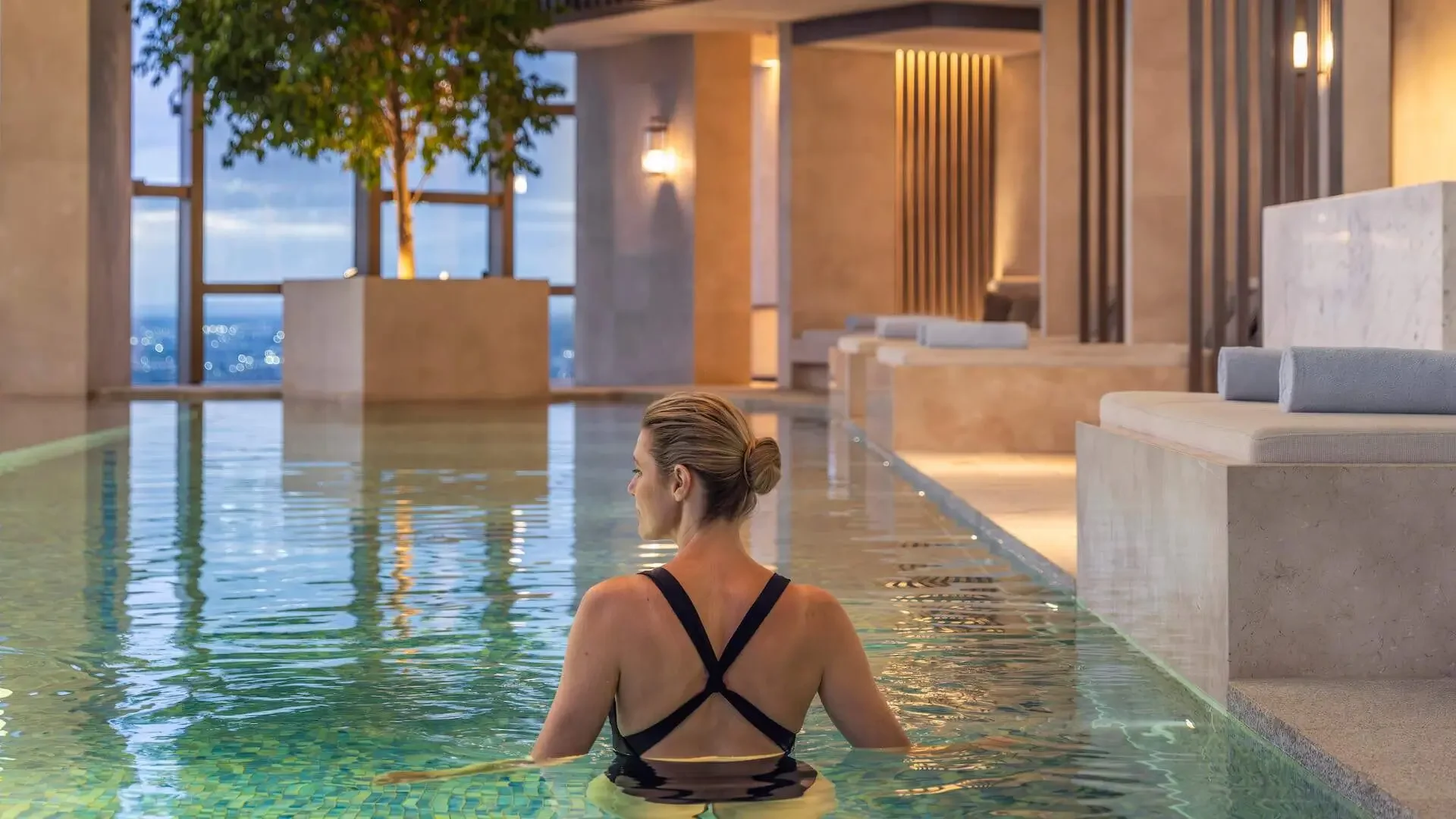 A woman with blonde hair tied in a bun is sitting in a swimming pool in a luxurious indoor spa or pool area with modern beige and wooden decor, large windows, and comfortable lounge chairs with rolled towels.