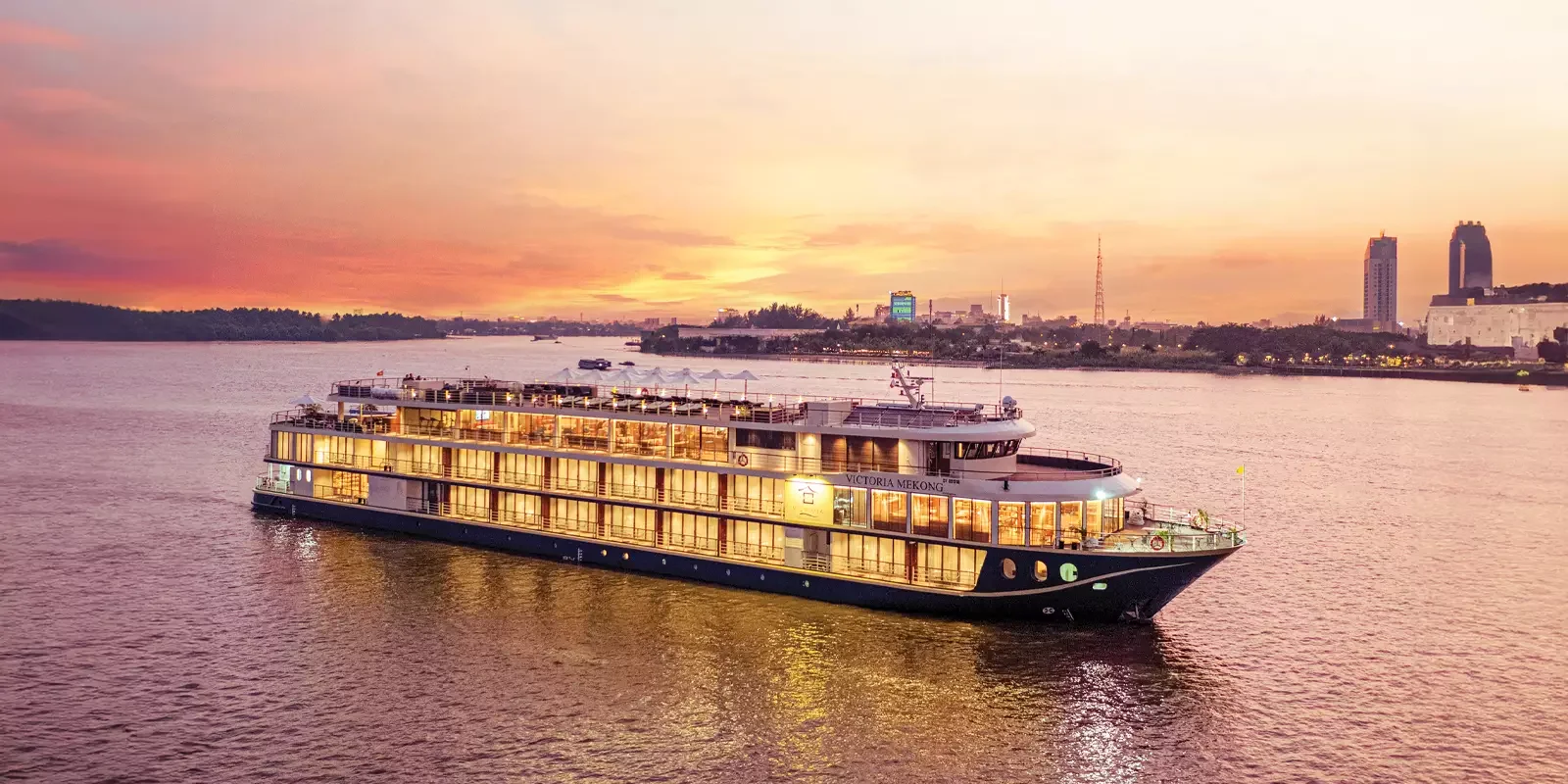A large cruise boat named Victoria Mekong sailing on a river during sunset, with illuminated windows and city skyline in the background.