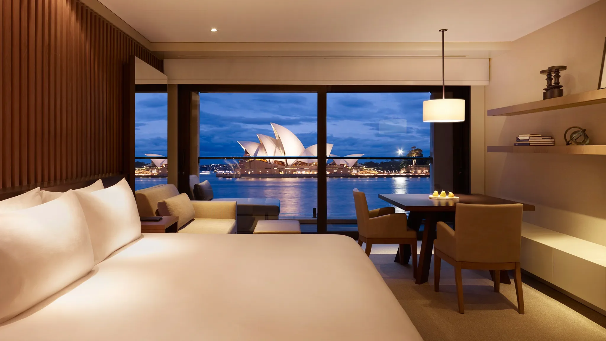 Hotel room with a view of the Sydney Opera House and harbor at night, featuring a bed, seating area, dining table, and open balcony.