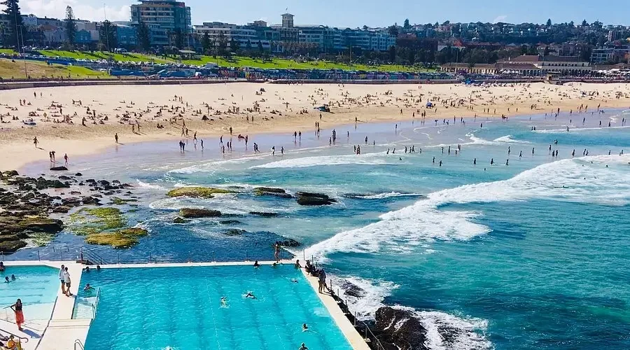 People relaxing and swimming at a beach with a sandy shoreline, rocky areas, and an ocean surf, with buildings in the background.
