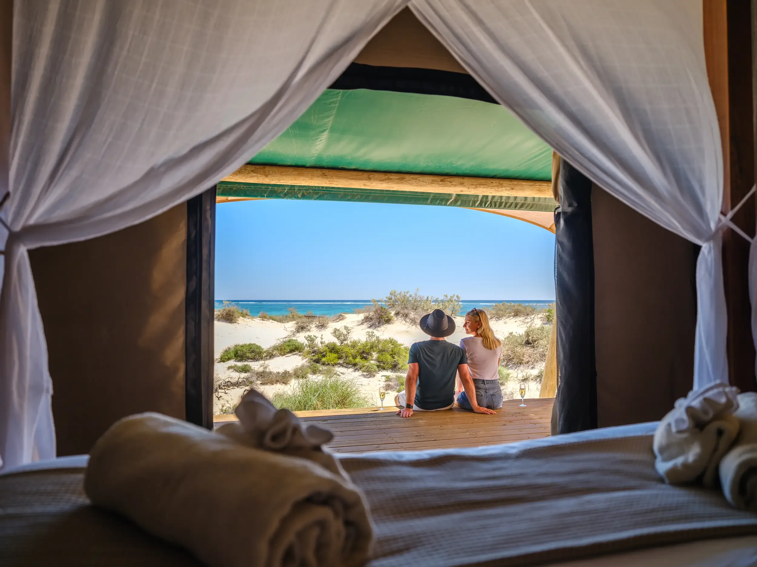 View from a tent looking out at two people sitting on a wooden deck near a sandy beach and ocean, with towels inside the tent in the foreground.