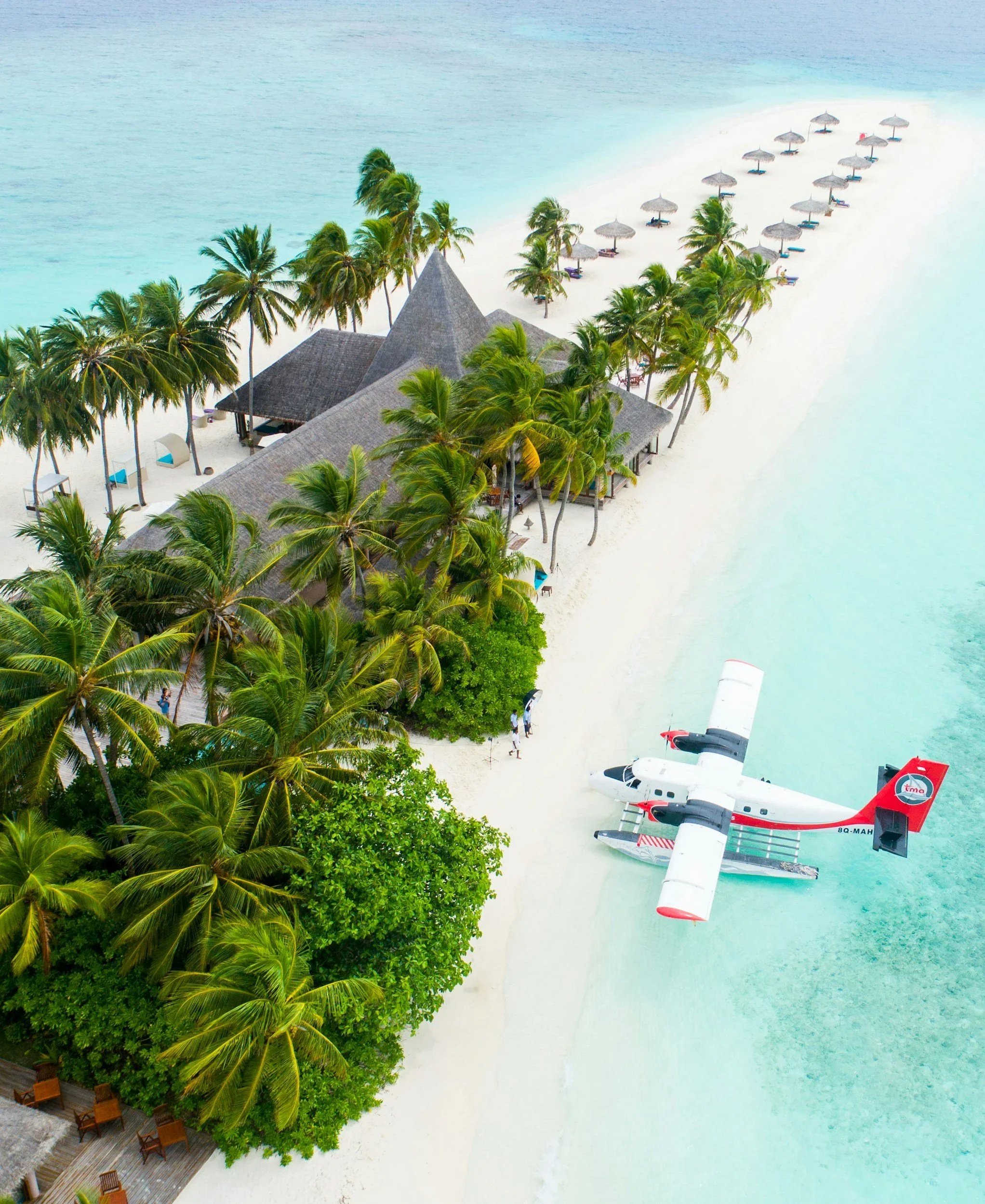 Aerial view of a tropical beach with a white sandy shore, turquoise water, palm trees, a thatched-roof building, beach umbrellas, lounge chairs, and a seaplane parked on the beach.