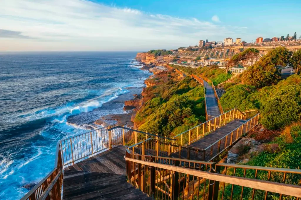A scenic coastal view with wooden walkways and railings on a hillside overlooking the ocean. There are homes on top of the hill and lush green foliage along the slope.