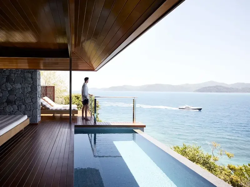 A person standing on a balcony overlooking the ocean, with a boat passing by in the water, and hills in the distance; part of a modern house with wooden flooring and a pool.