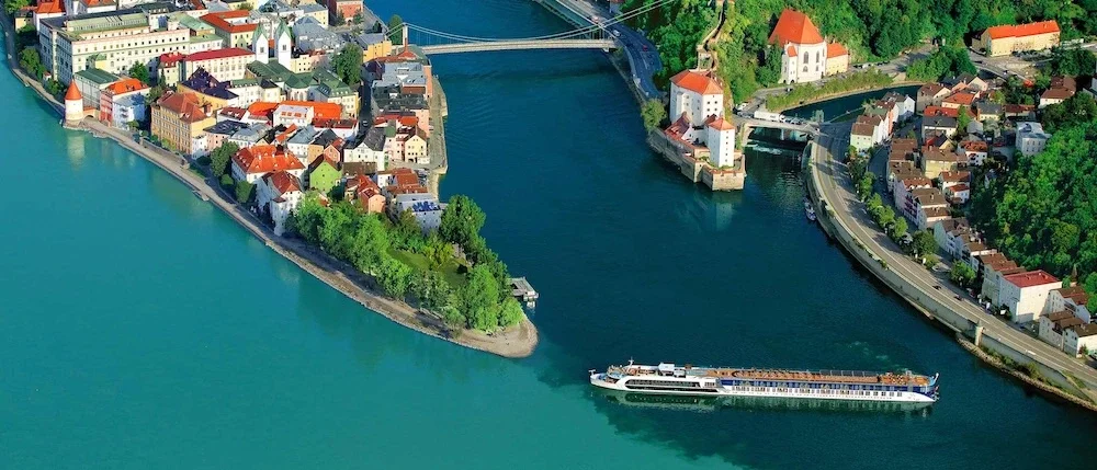 Aerial view of a European city with a river, historic buildings with red roofs, and a large white cruise ship sailing on the river.