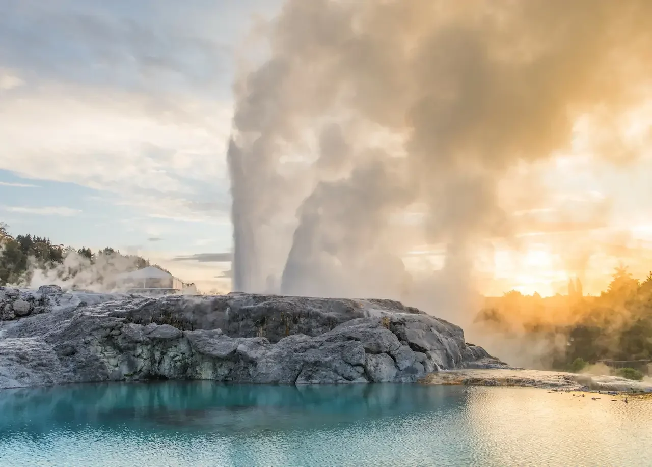 Geothermal geyser erupting with steam and water, surrounded by a hot spring pool at sunset.