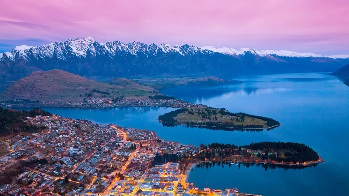 Aerial view of a lakeside town with mountains in the background during sunset, featuring snow-capped peaks and calm waters.