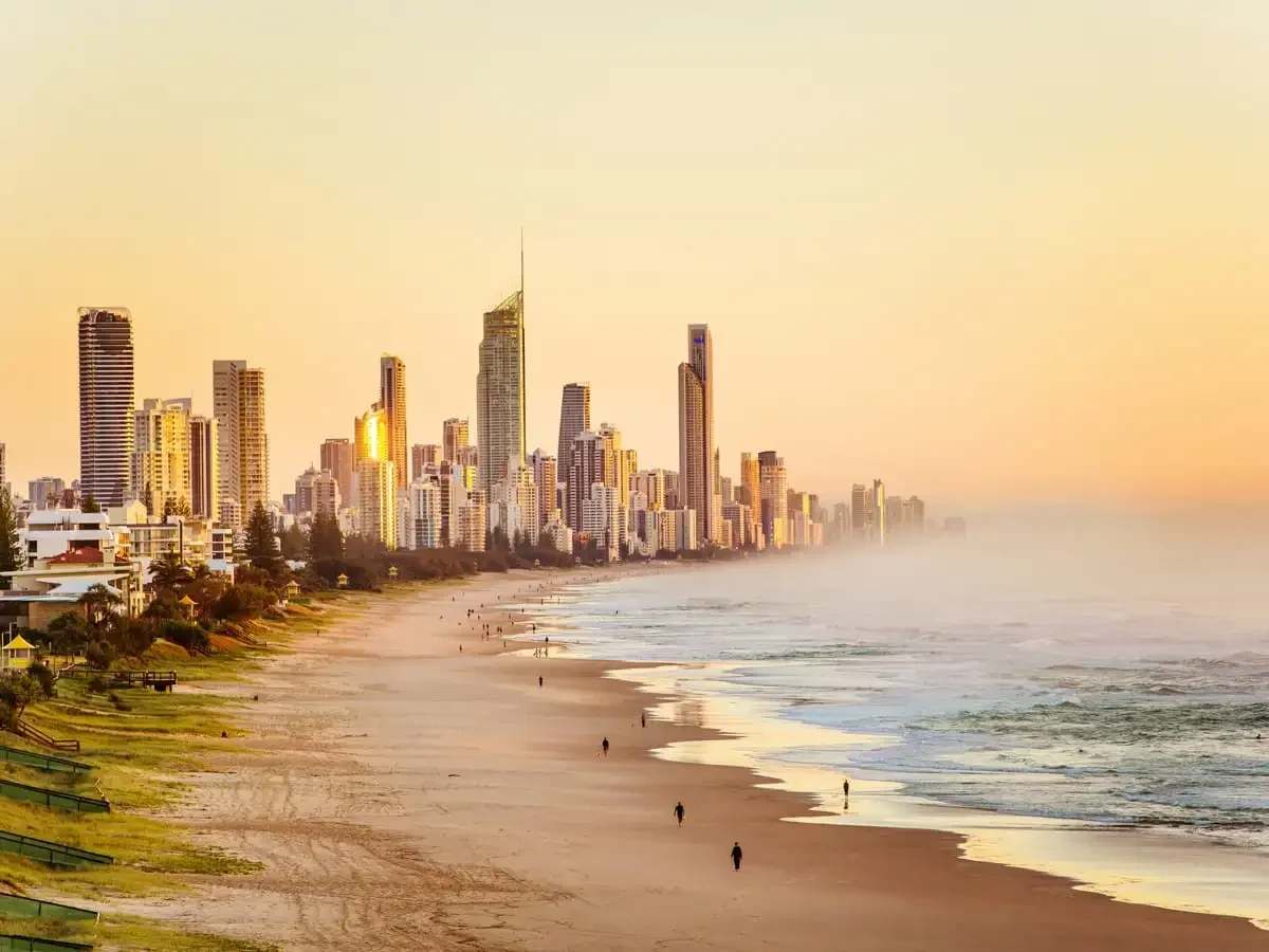 City skyline with tall skyscrapers and a beach with a few people walking along the shoreline during sunset or sunrise.