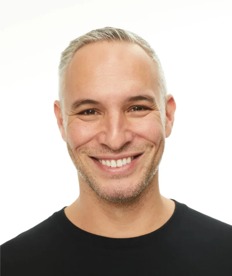 A smiling man with short, gray hair, slight stubble, wearing a black shirt, against a white background.