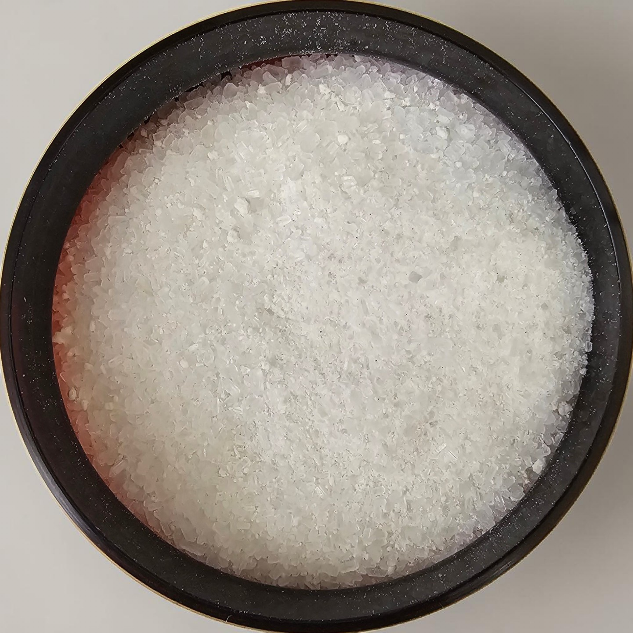 Close-up of a black circular container filled with coarse white sea salt on a white surface.