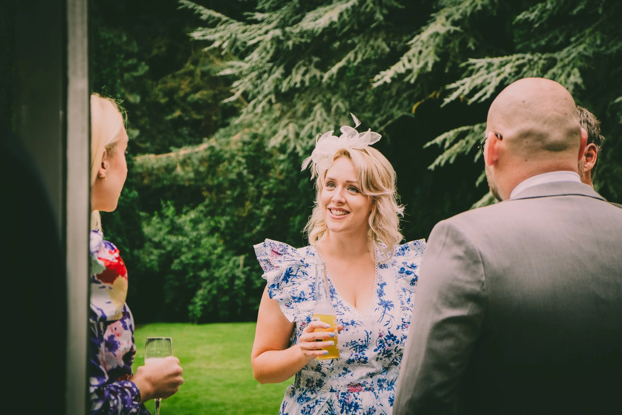 A group of people socializing outdoors in a garden setting. One woman in the center is smiling, holding a drink, and wearing a white floral dress with a decorative fascinator. Others are partially visible, holding drinks, and engaged in conversation.