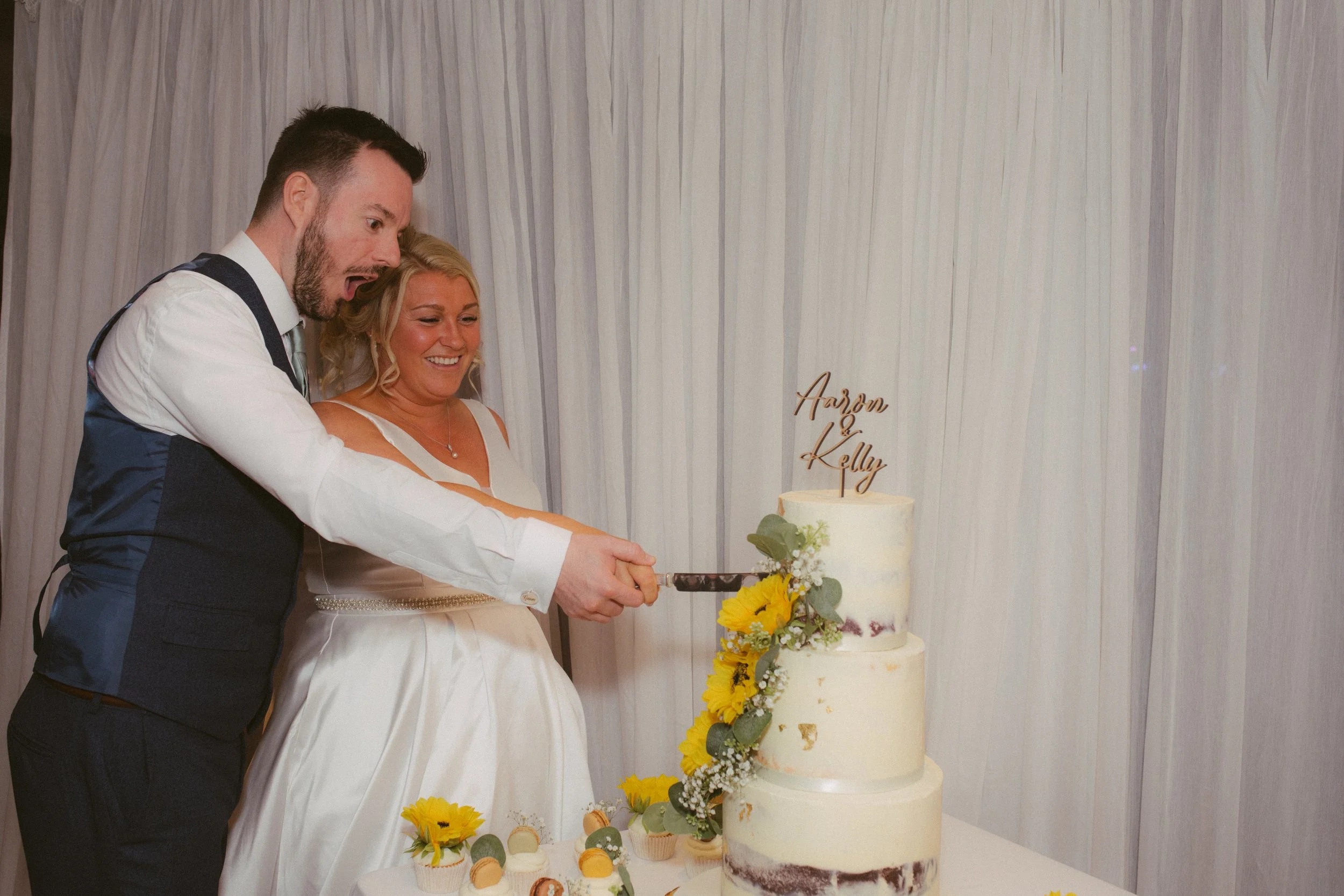 A newlywed couple cutting a wedding cake, with a backdrop featuring their names 'Harson & Kelly'.