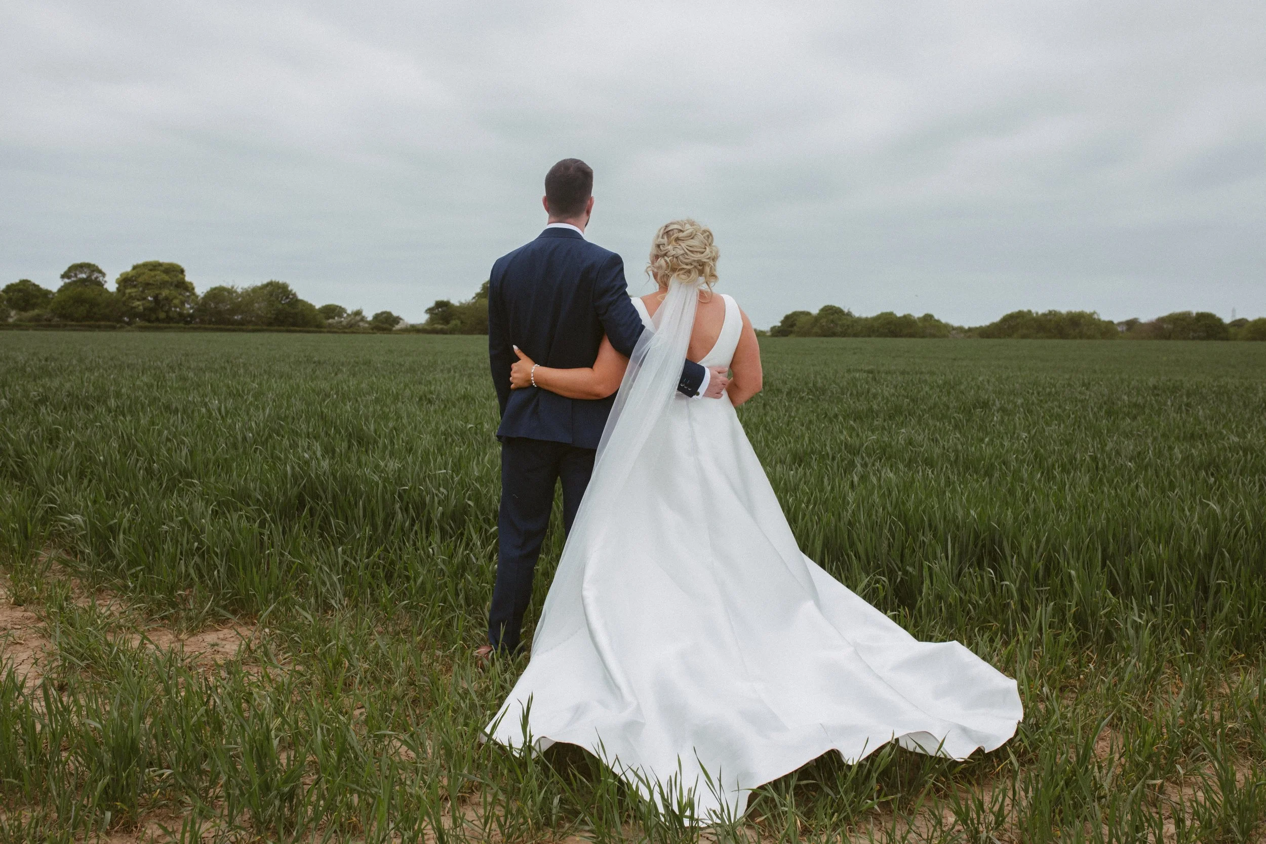 A bride and groom stand together in a green field looking towards a cloudy sky, with the bride's white wedding dress flowing behind her.