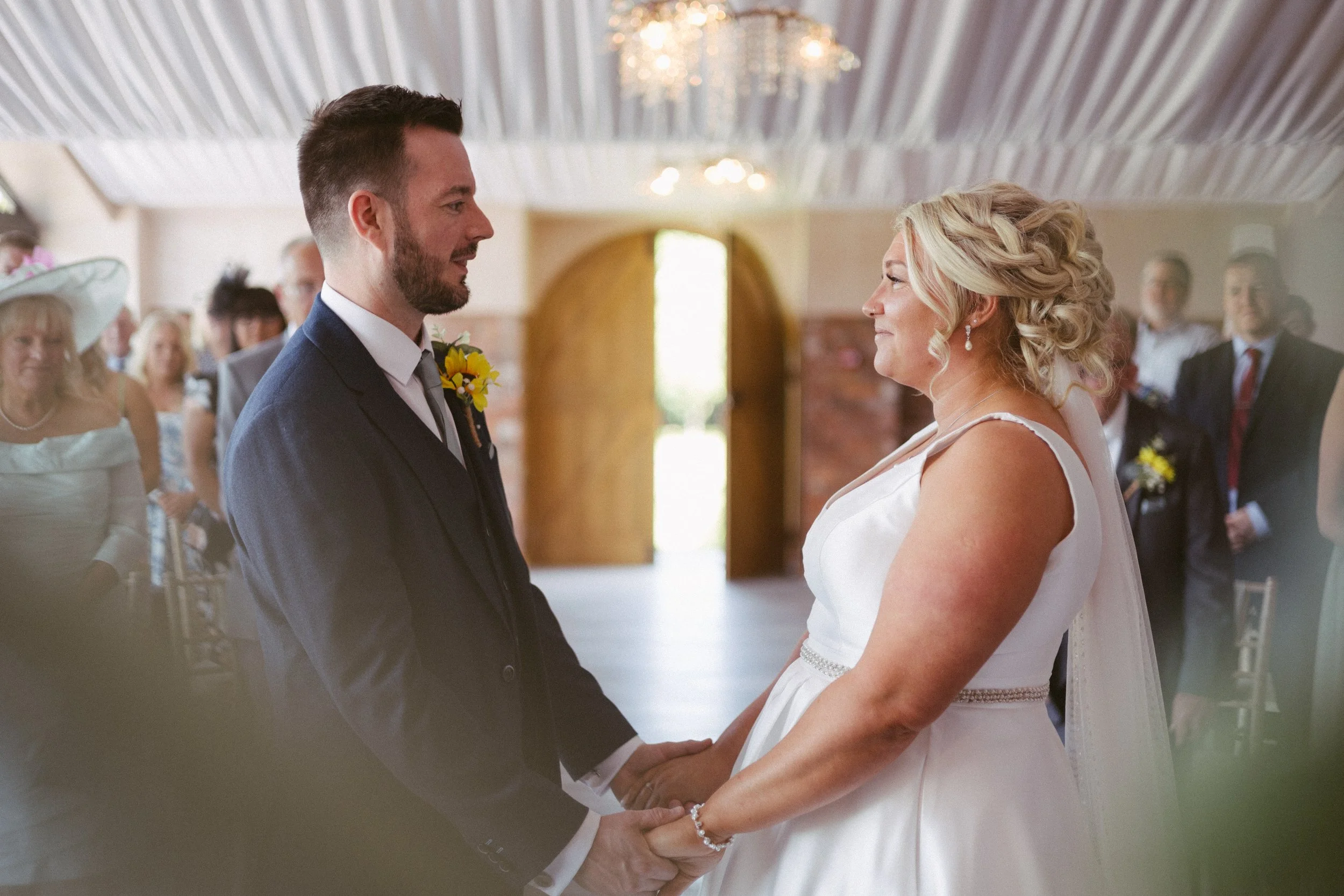 A bride and groom holding hands and looking at each other during their wedding ceremony indoors, with guests watching in the background.