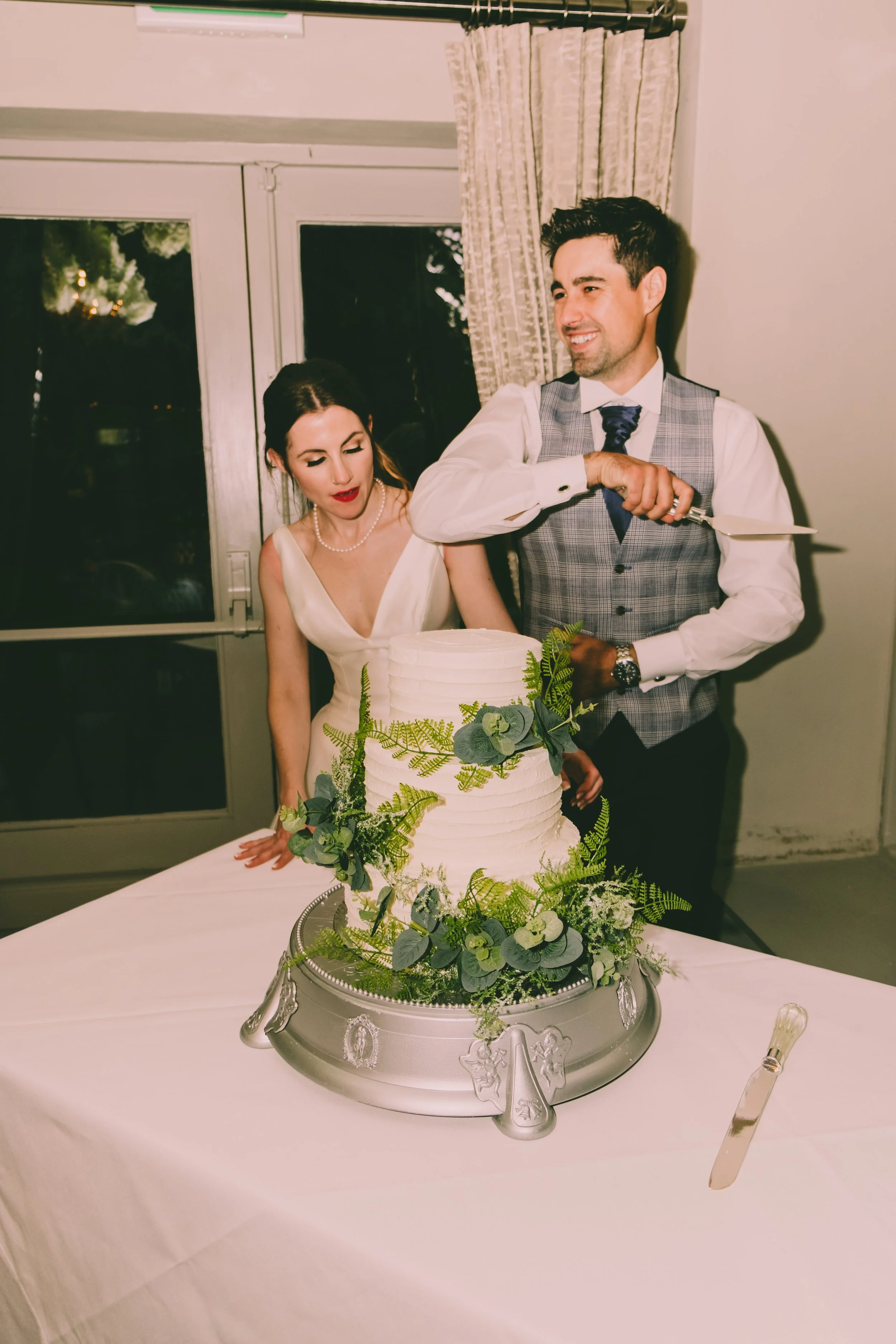 A man and a woman at a wedding celebration, the man is smiling and using a cake knife to cut a three-tier wedding cake decorated with greenery. The woman is wearing a white dress, and the man is wearing a gray vest and tie. The cake is on a silver pl