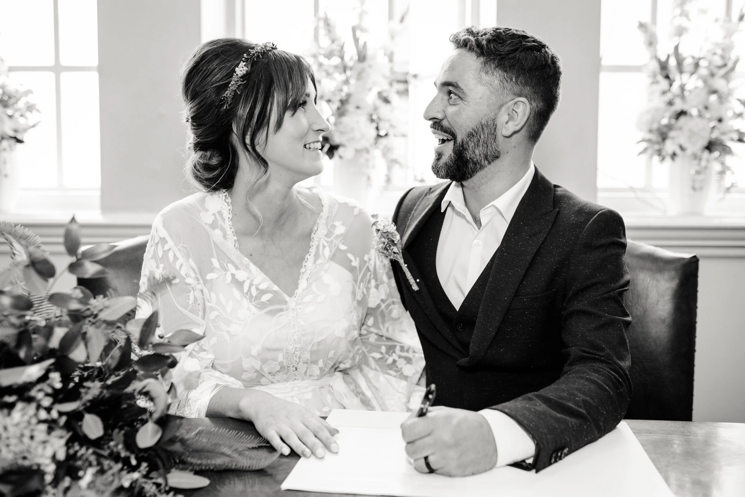 A bride and groom smiling at each other during their wedding ceremony, seated at a table with greenery and flowers in the foreground, in a bright room with large windows.