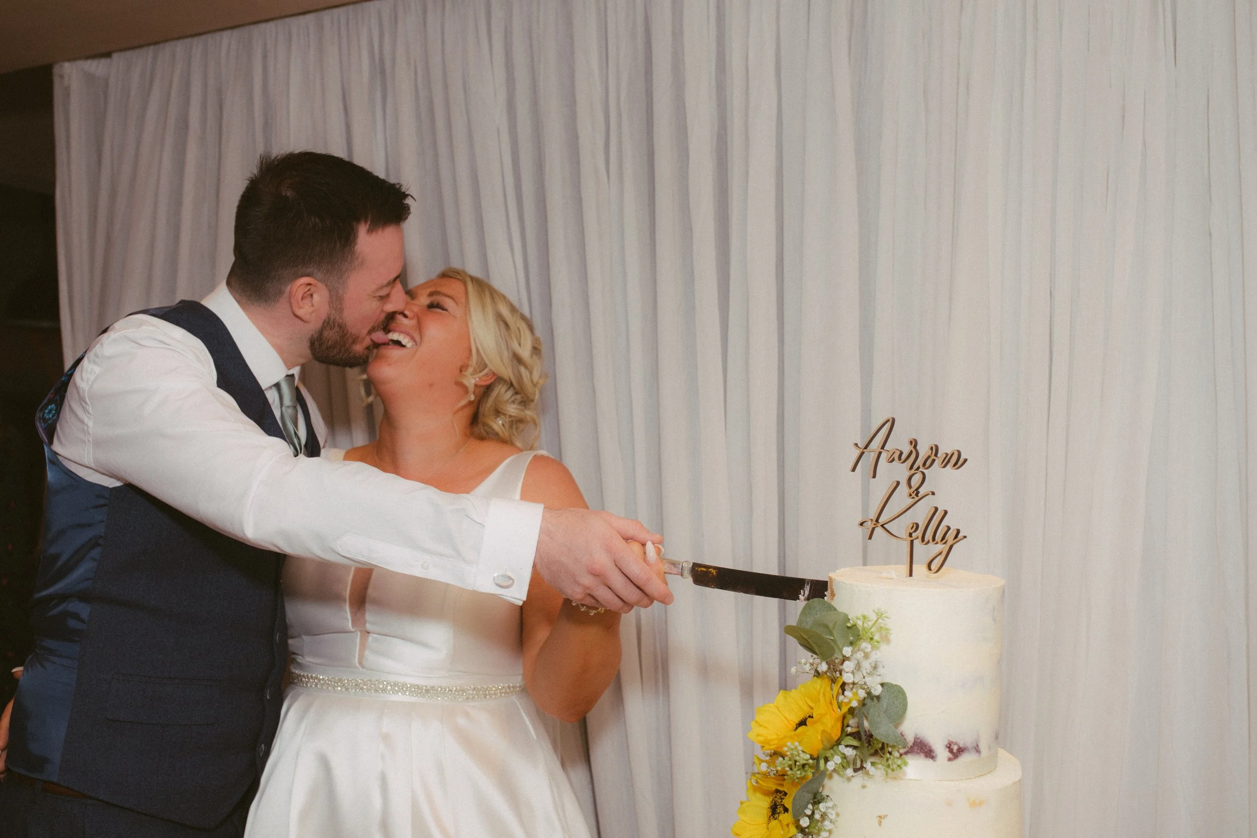 A couple in wedding attire is cutting a wedding cake together; the cake has a topper with the names Aaron and Kelly, decorated with yellow and white flowers, in front of a white curtain backdrop.