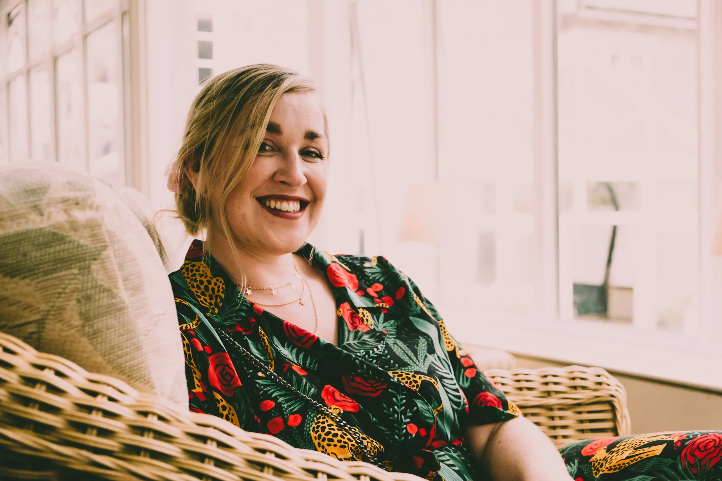 A woman sitting on a wicker chair, smiling, with sunlight coming through large windows in a bright room.