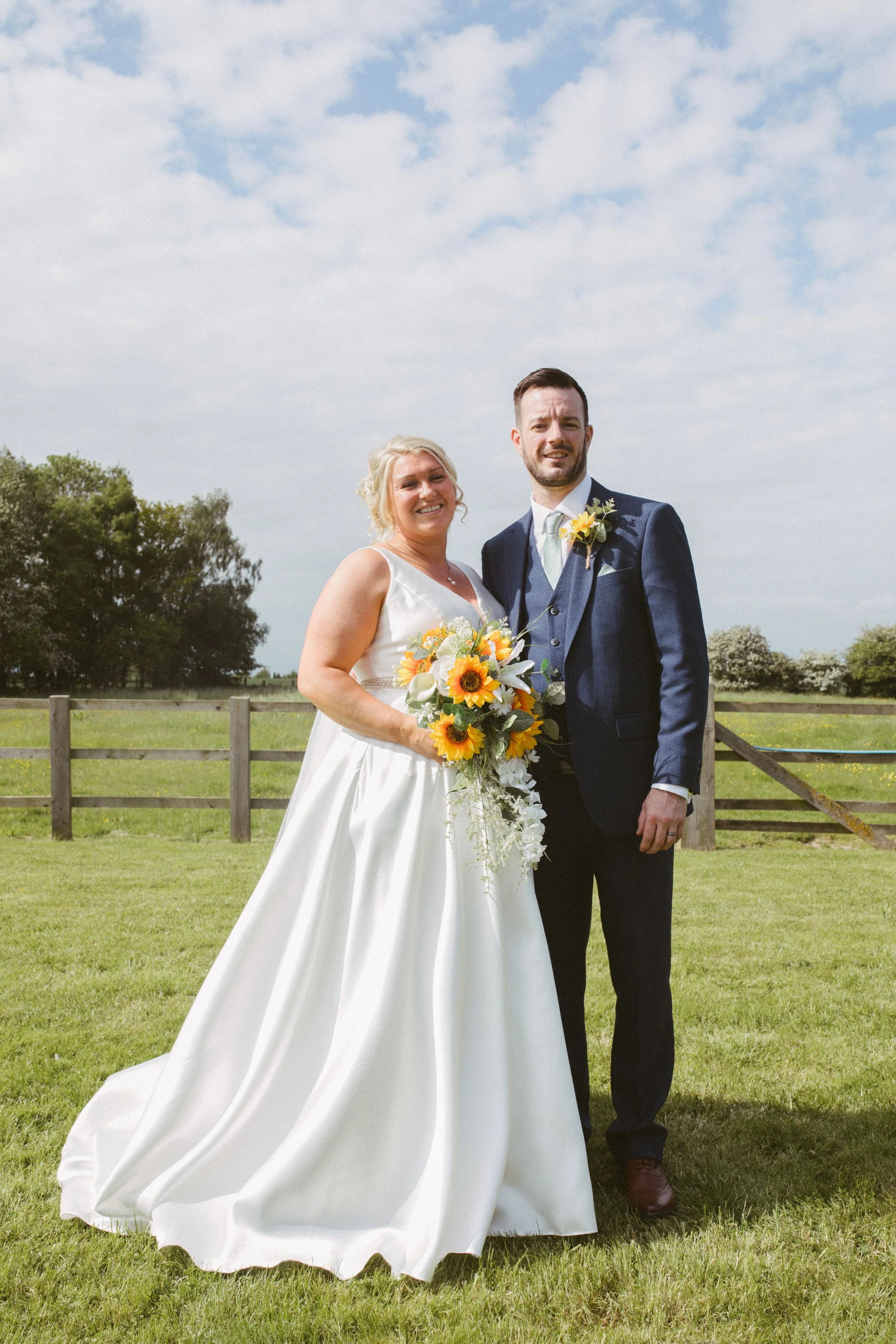 A bride and groom standing outdoors on a grassy field, smiling at the camera. The bride is holding a bouquet of sunflowers and white flowers, wearing a white wedding gown. The groom is wearing a dark suit with a white shirt and light-colored tie. The