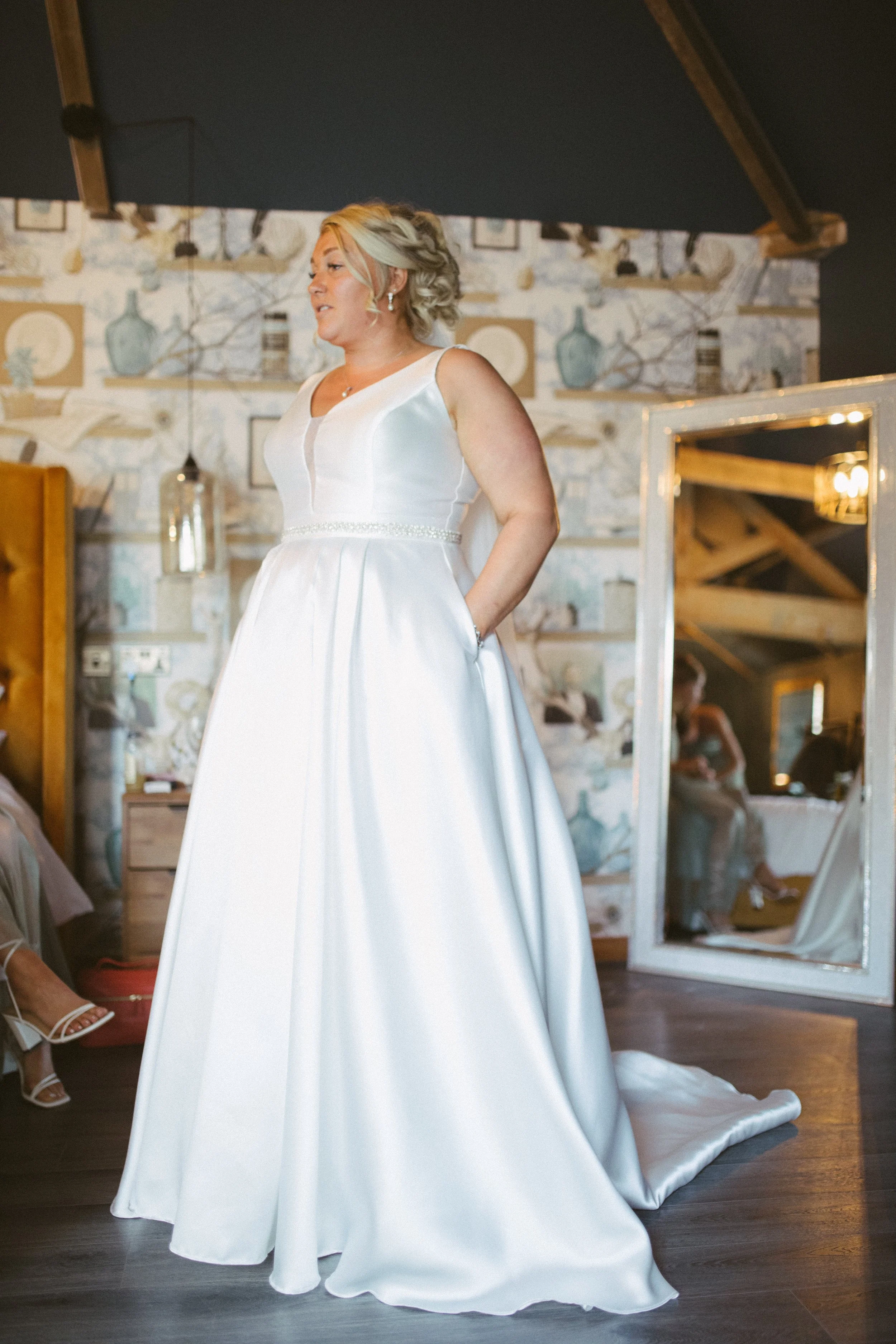 A woman in a white wedding dress standing in a rustic room with exposed brick walls and wooden beams, reflected in a large mirror.