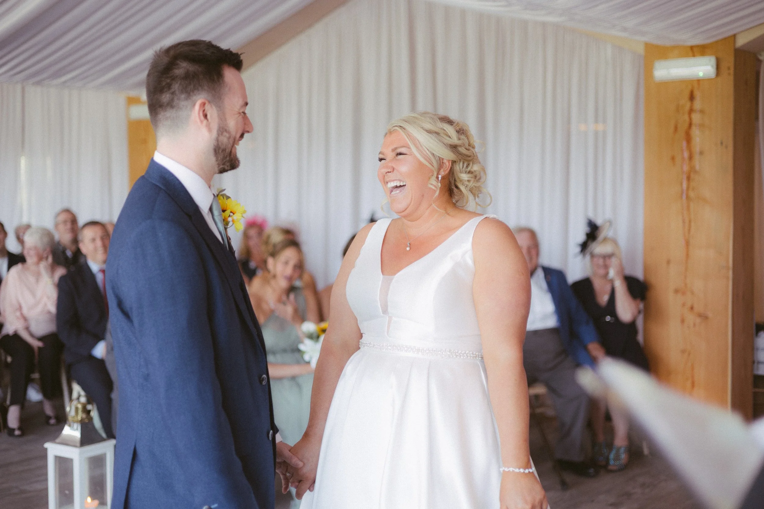 A bride and groom holding hands during a wedding ceremony, smiling at each other, with guests in the background.