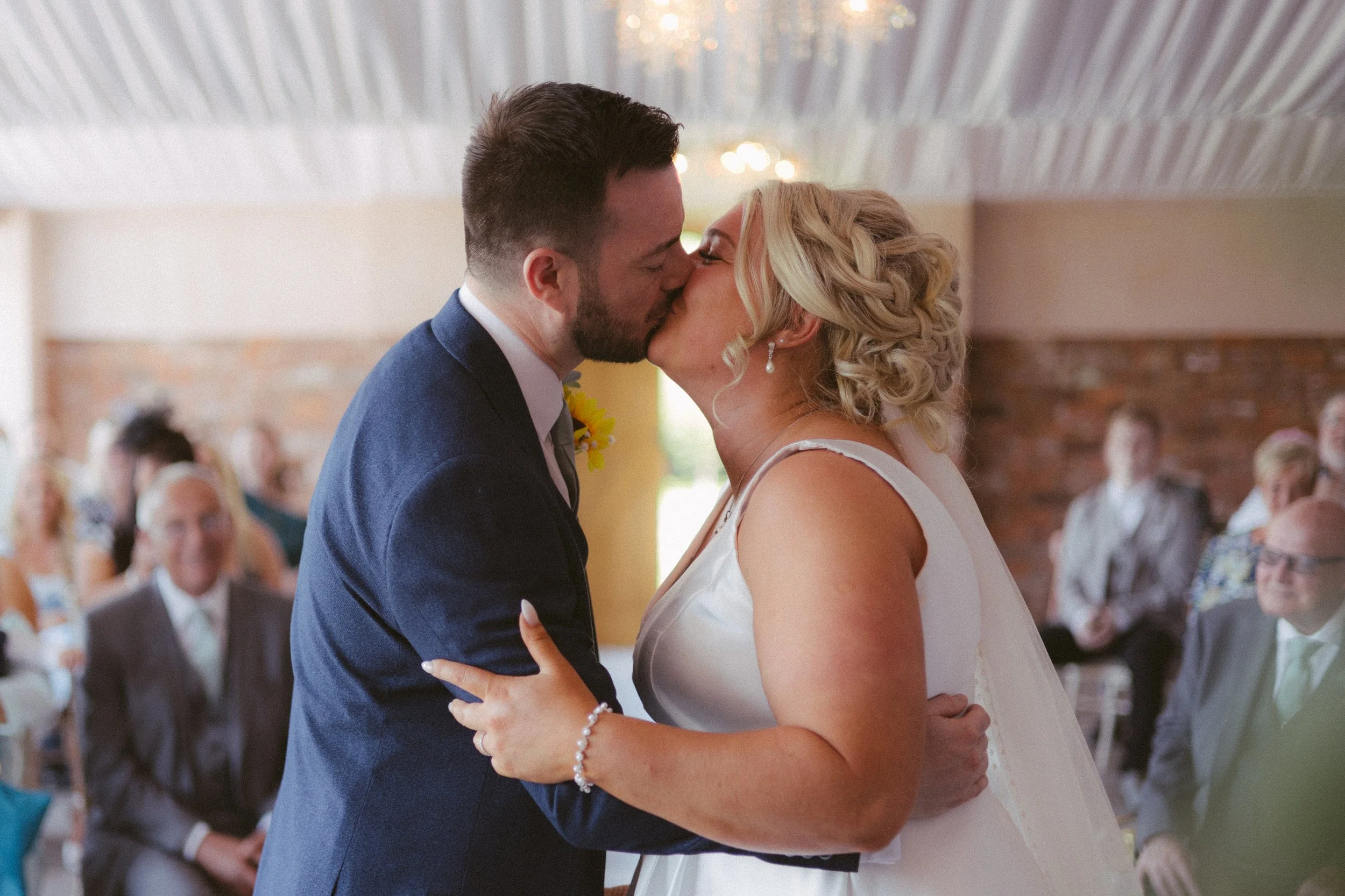 A newlywed couple sharing a kiss during their wedding ceremony, with guests seated in the background.