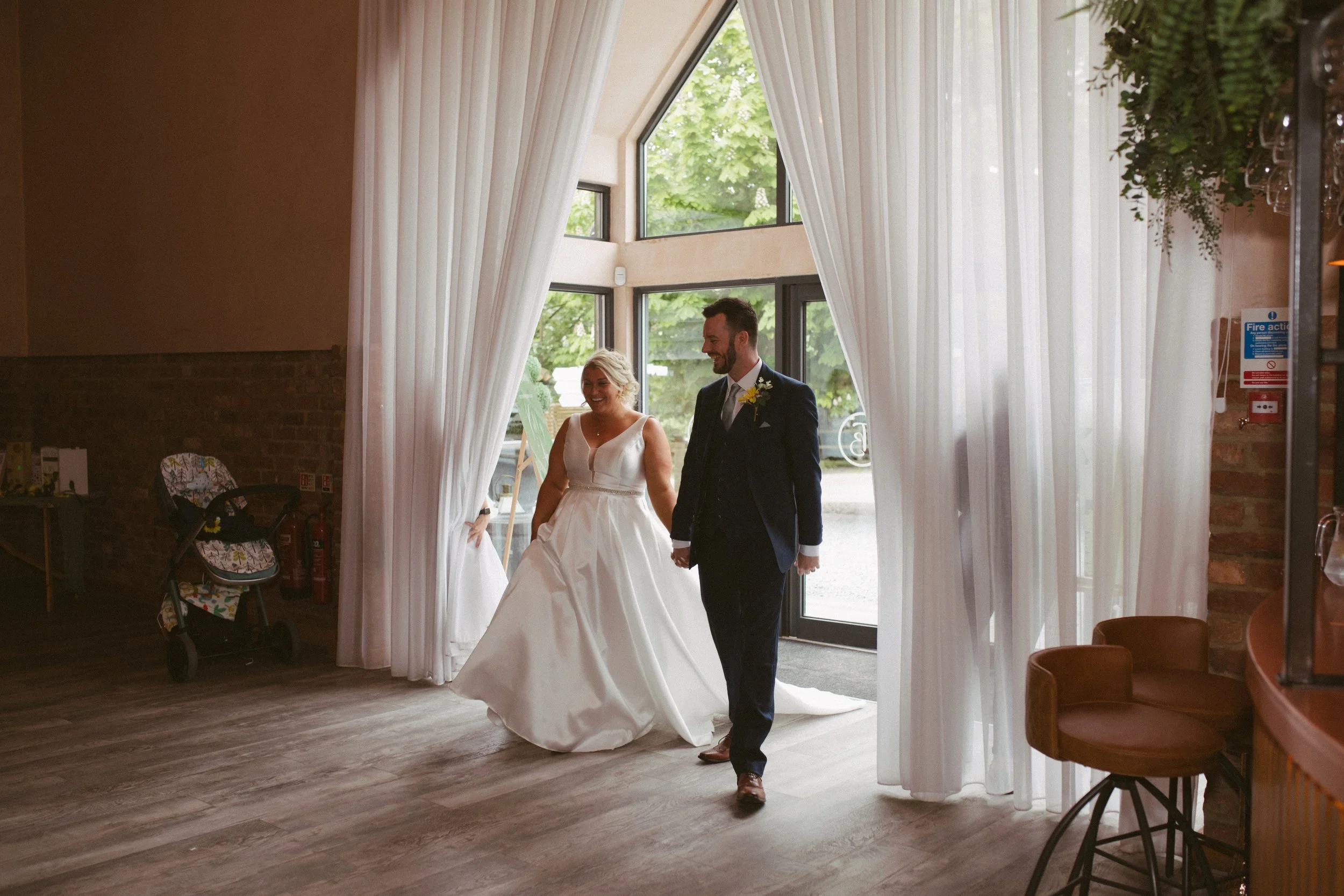 A bride and groom entering a venue through large white curtains, smiling and holding hands. The bride wears a white wedding dress, and the groom is in a dark suit with a boutonniere. The background shows green trees outside the venue.