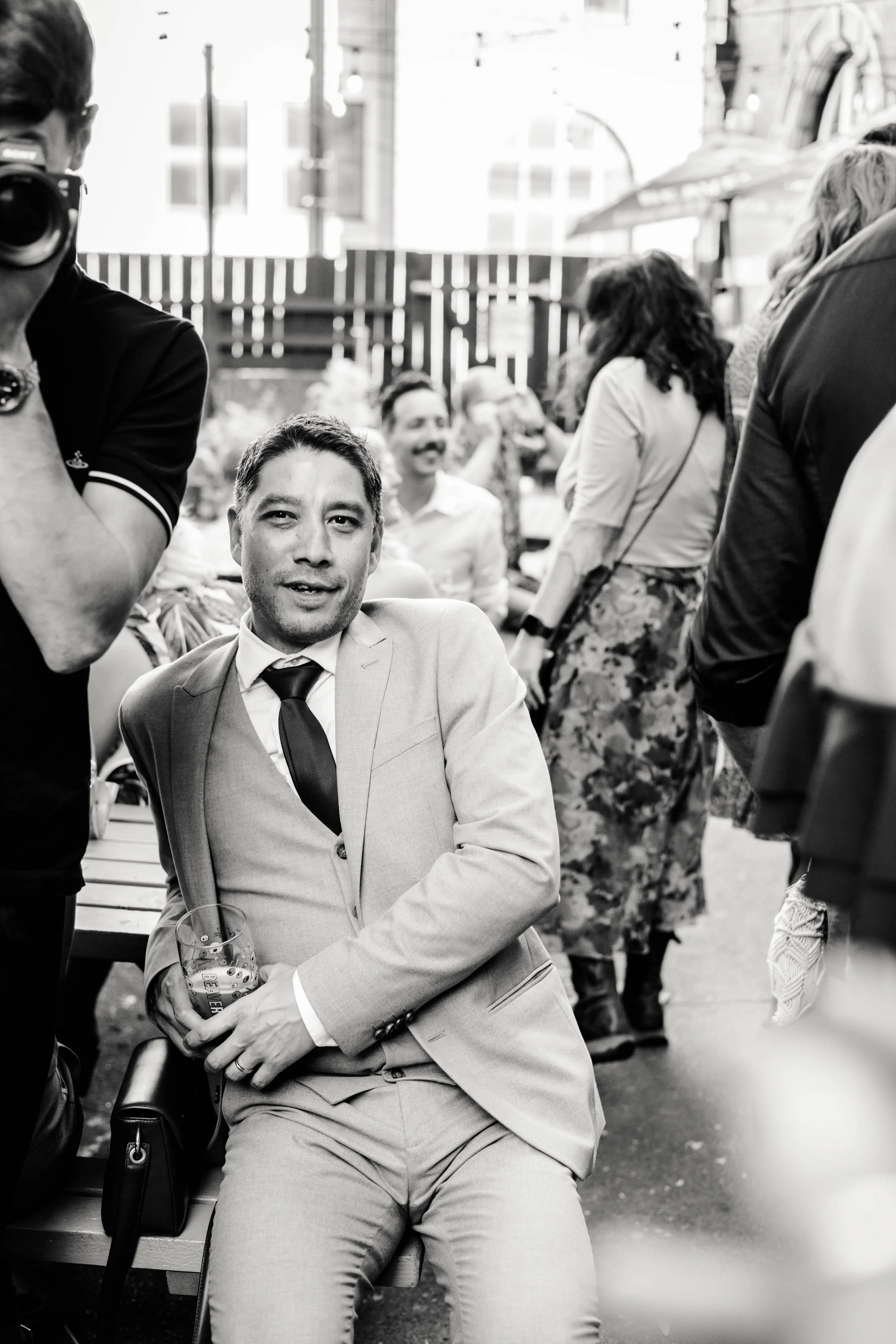 A black-and-white photo of a man in a light suit and tie sitting on a bench, holding a glass of beer, with other people socializing in the background at an outdoor gathering.