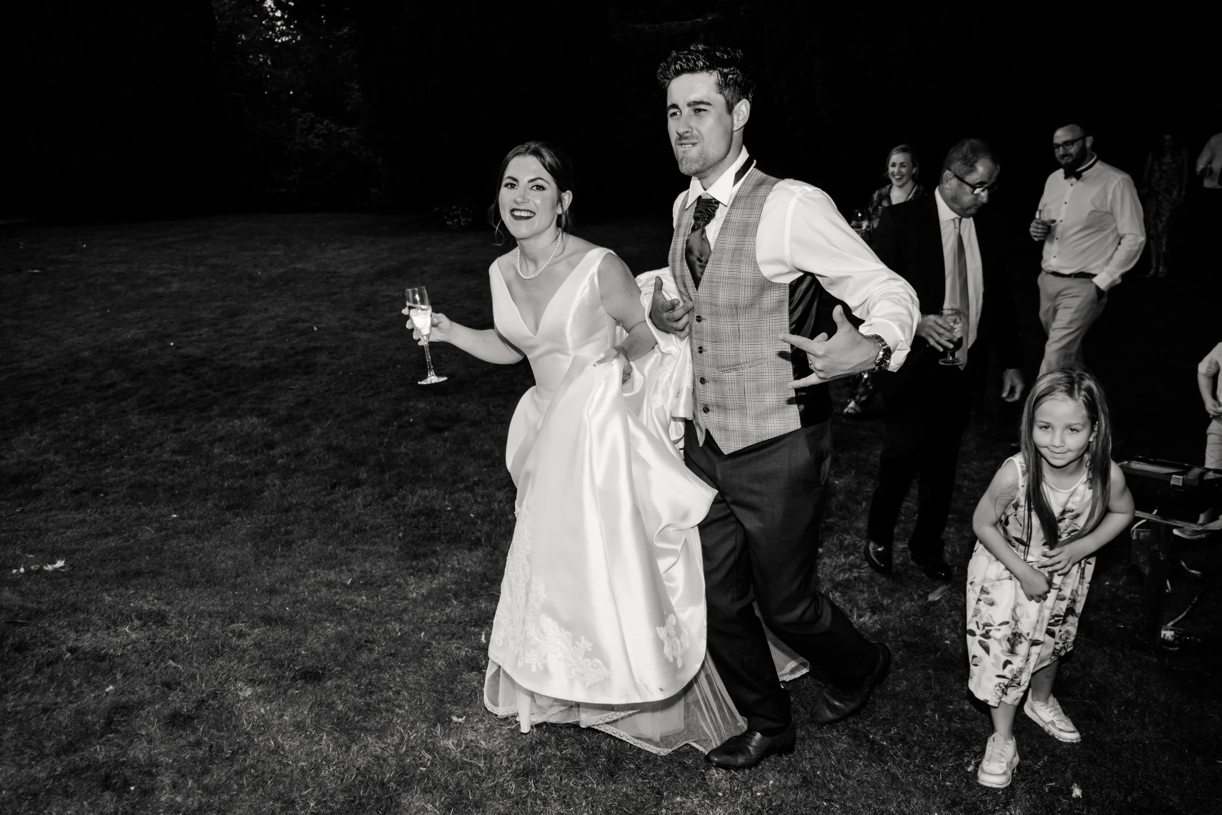 A black and white photo of a bride and groom at a wedding reception outside at night. The bride is holding a champagne glass and smiling, while the groom is looking away with a playful gesture. Several other guests are in the background, some holding