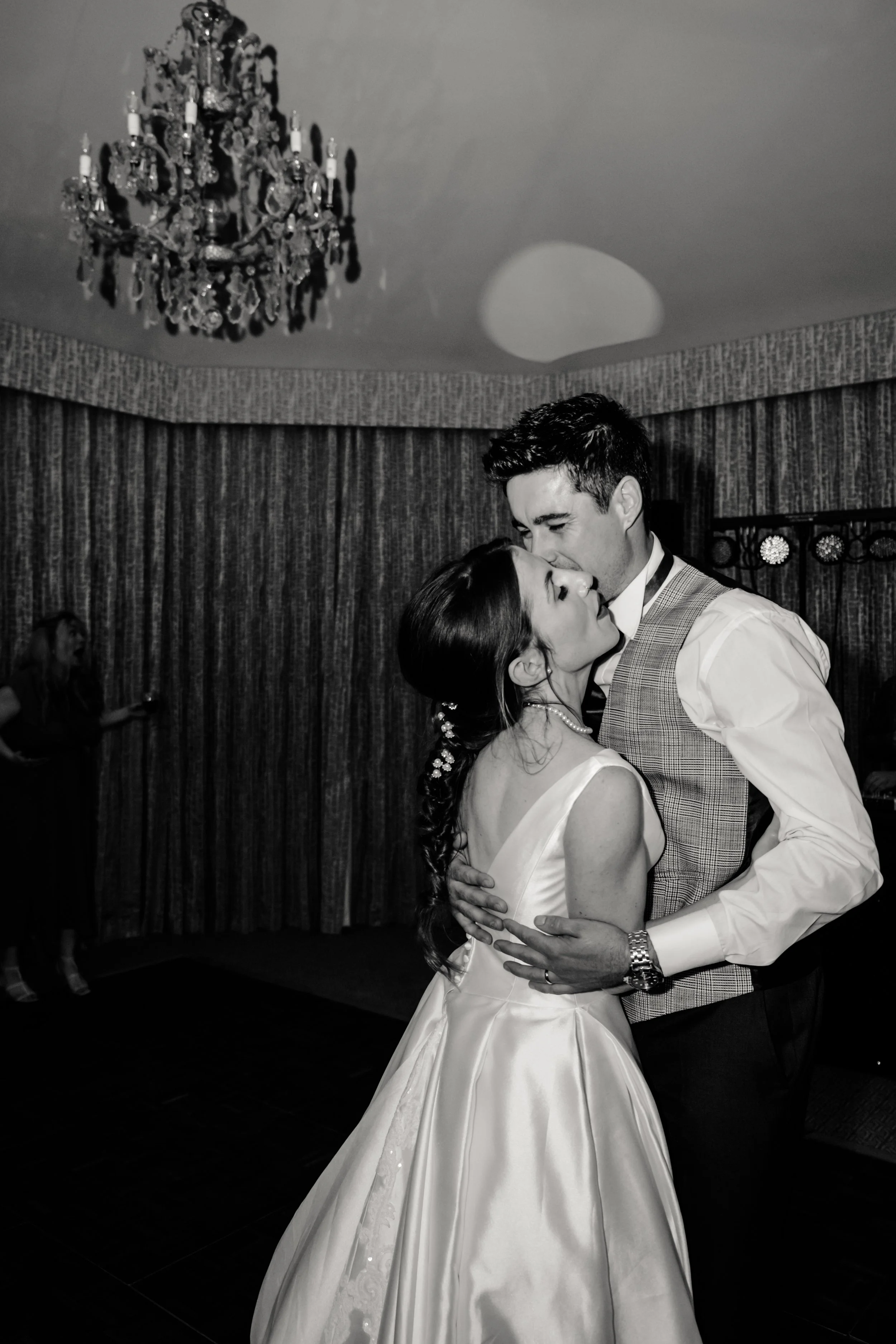 A black-and-white photo of a couple dancing closely at a formal event or wedding reception. The woman is wearing a satin wedding dress and pearl necklace, and the man is dressed in a collared shirt and vest. The background features a textured curtain