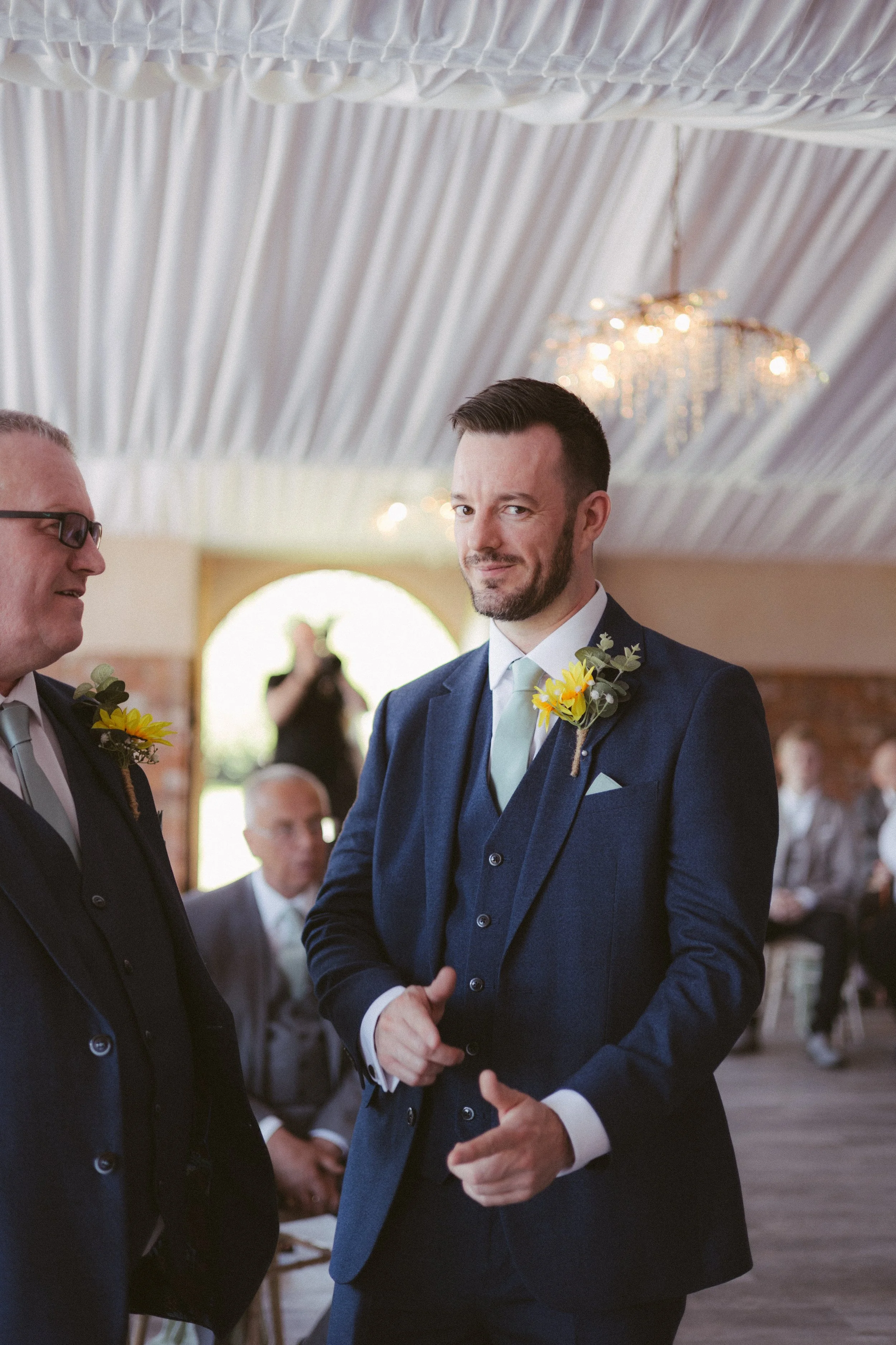A groom in a dark blue suit with a sunflower boutonniere contrasts with the white and elegant attire of the other guests at a wedding ceremony in a decorated hall with a crystal chandelier.
