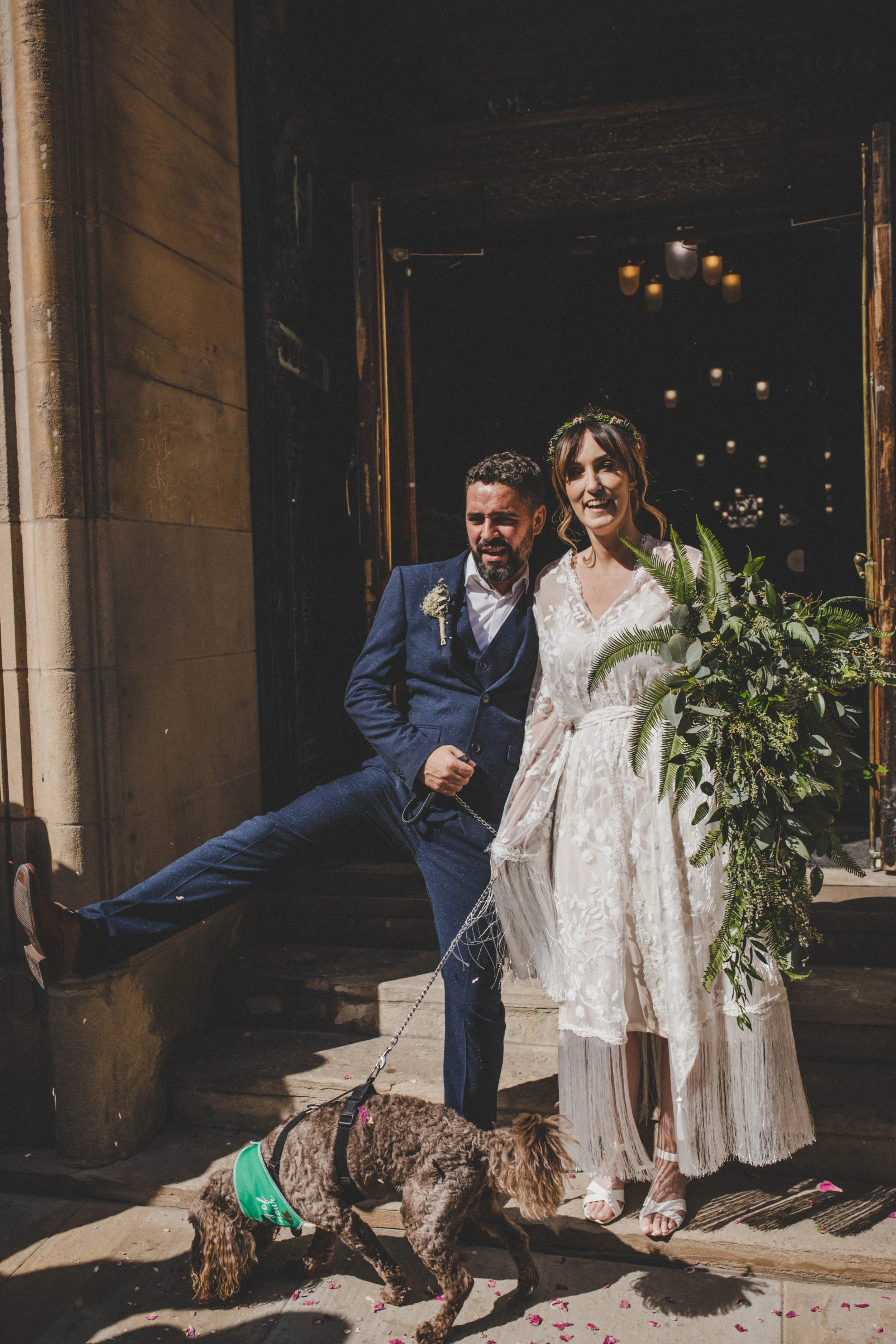A couple, dressed as a bride and groom, stands outside a building with a dog in front. The groom is in a navy blue suit and the bride is in a white lace dress with a bouquet, both smiling. The dog, a brown and black spaniel, wears a green vest and is