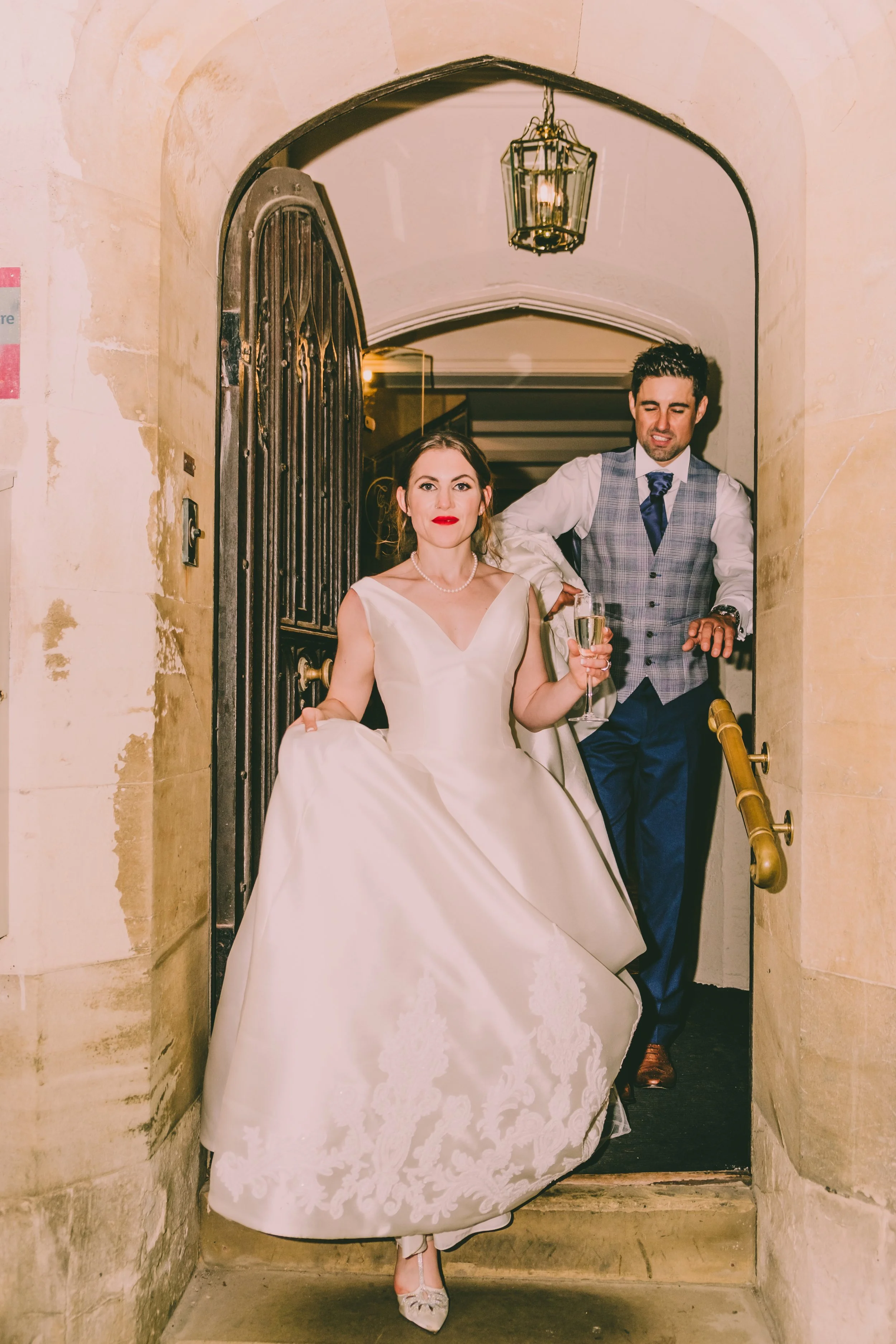 A bride in a white gown holding a glass of champagne, standing in a stone archway, with a man in a vest and tie behind her.