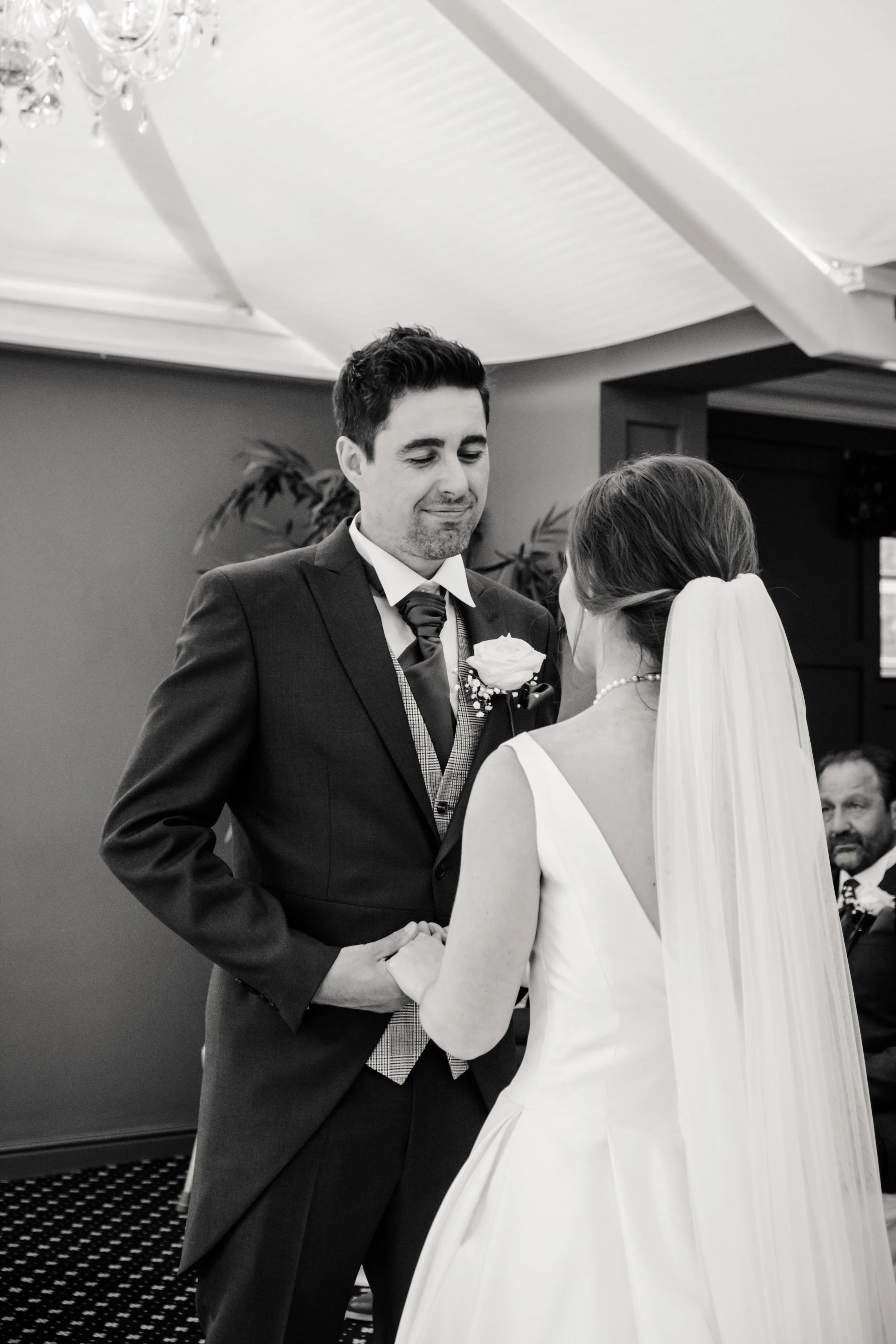 A groom and bride holding hands during their wedding ceremony, with the groom smiling and the bride's face partially visible, inside a decorated venue.