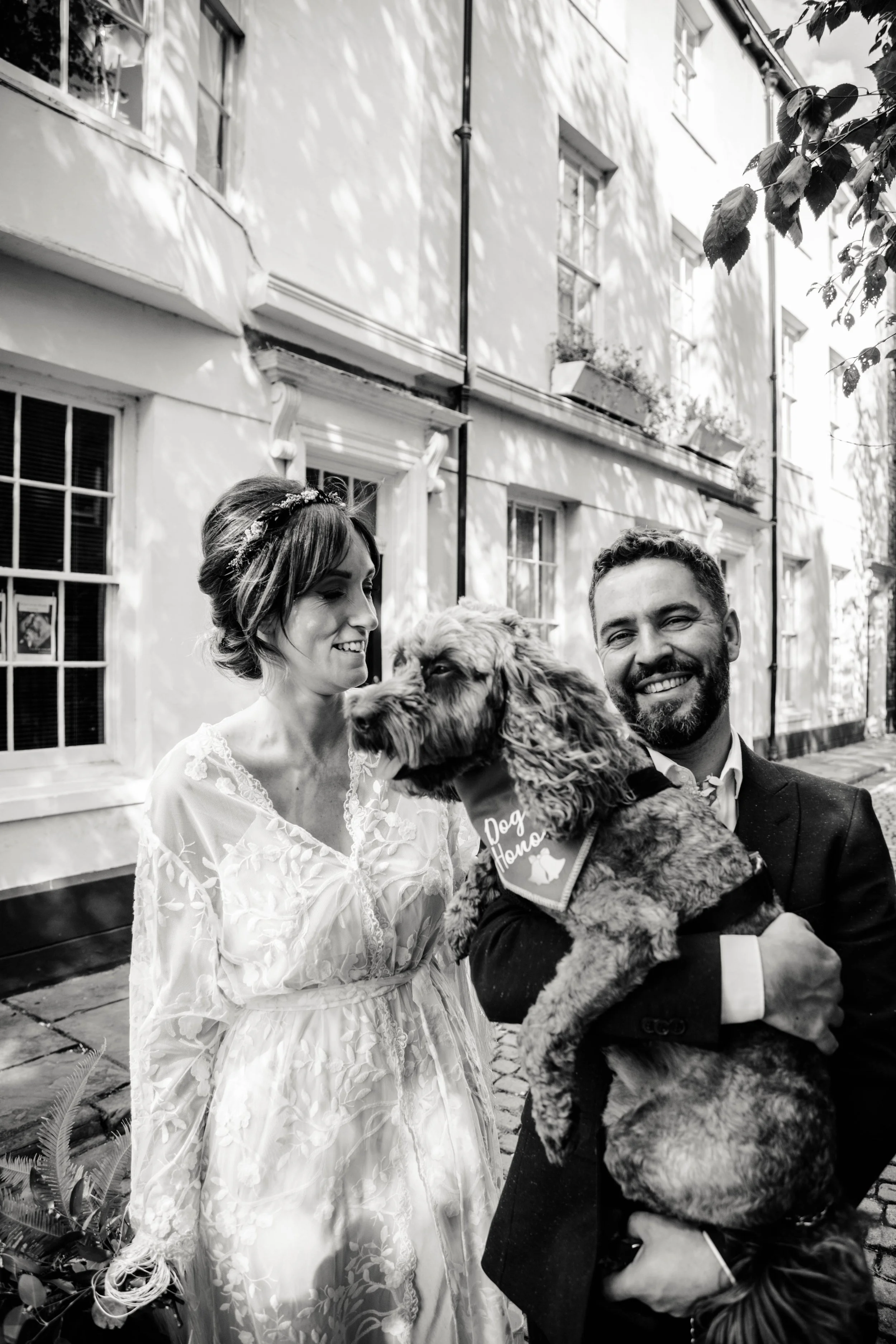 A black and white photo of a bride and groom outdoors on a wedding day. The bride, wearing a wedding dress with lace details and a floral headband, looks at a large dog. The groom, smiling, holds the dog, which has a bandana that says 'Dog Hona' and 