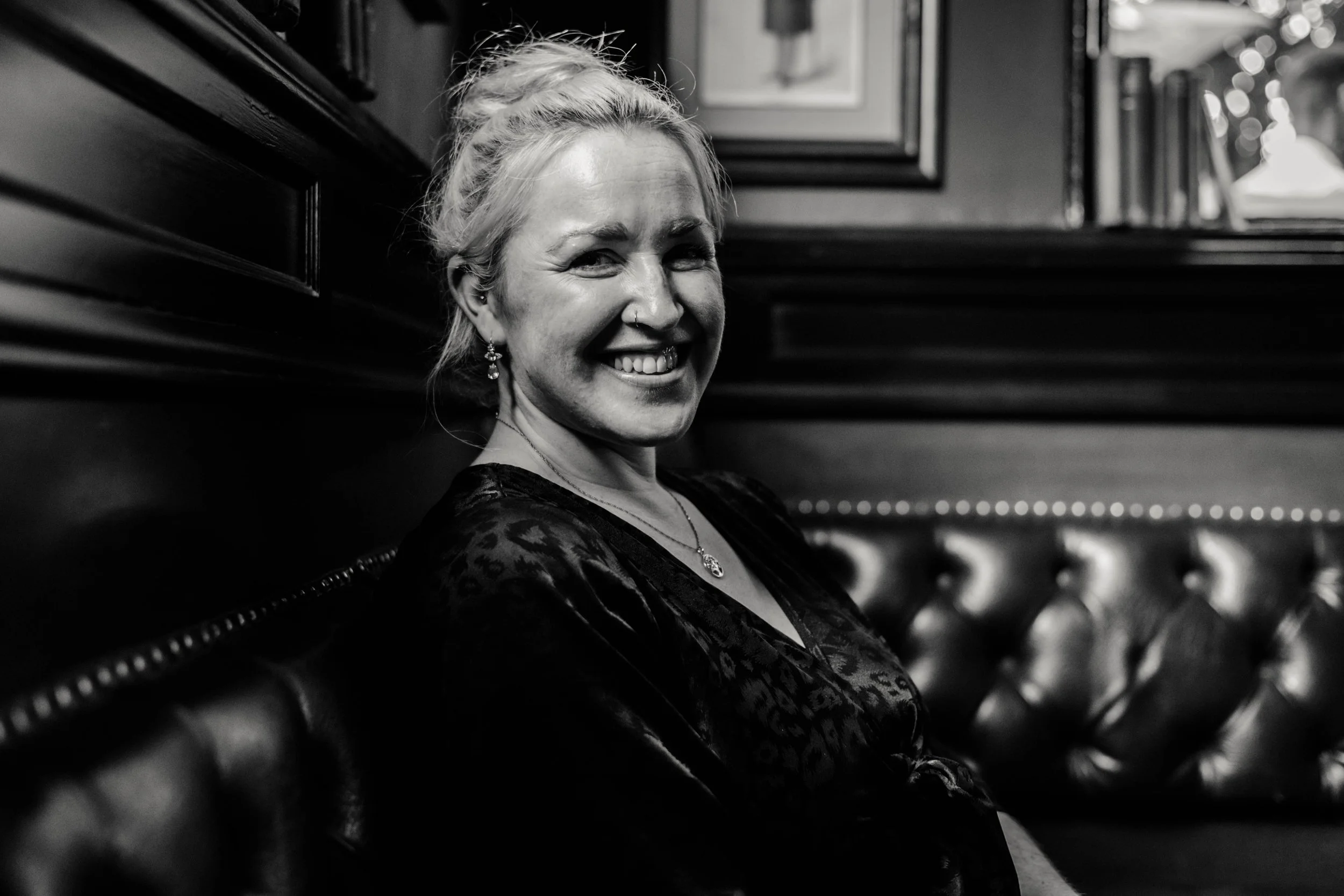 A woman with blonde hair in a bun smiling at the camera, sitting on a leather bench with framed pictures on the wall behind her, in black and white.