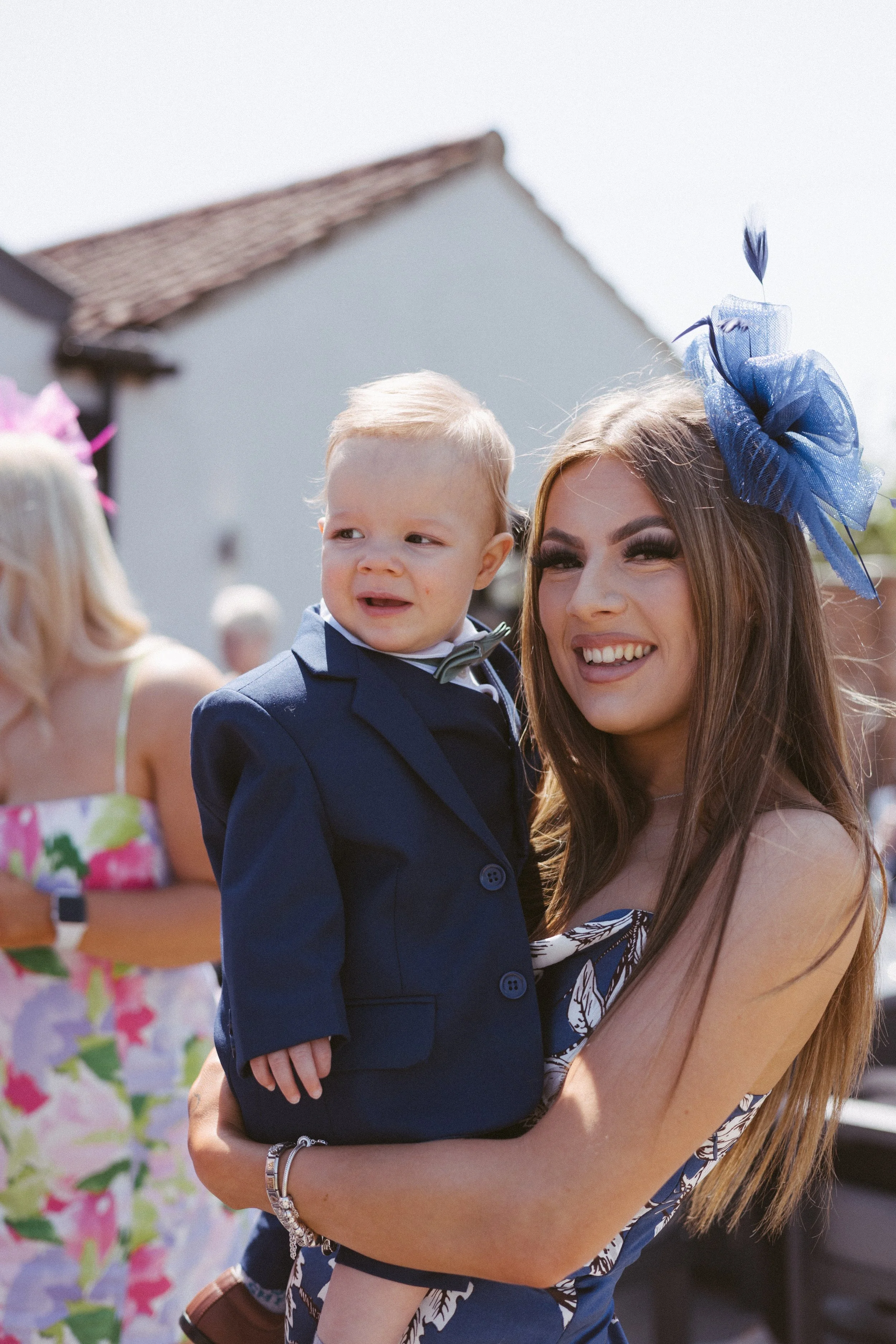 A woman with long brown hair and a large blue fascinator hat holding a young boy in a navy blue suit with a bow tie at an outdoor event with other people in the background.