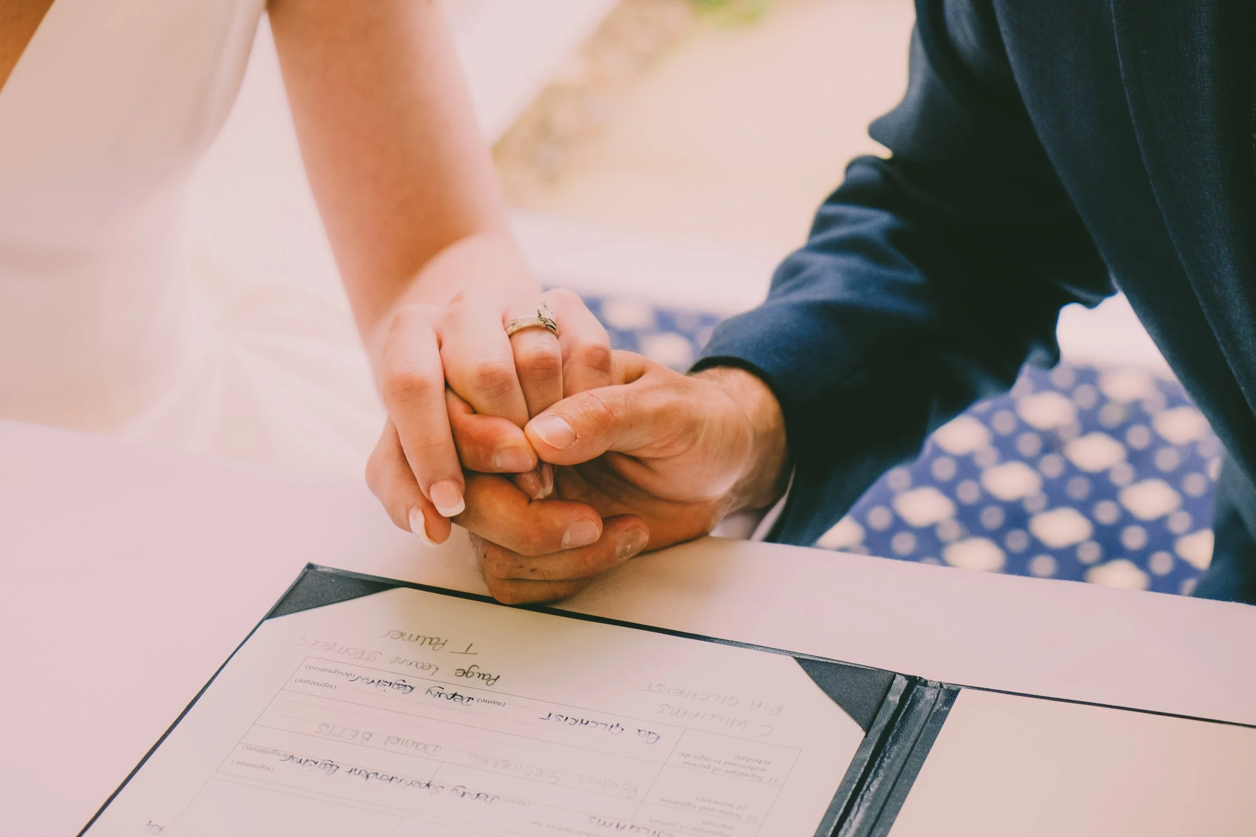 A person signing a document at a desk, with a handwritten wedding plan on the paper. The person is wearing a dark jacket and holding hands with another person, who is wearing a white dress.