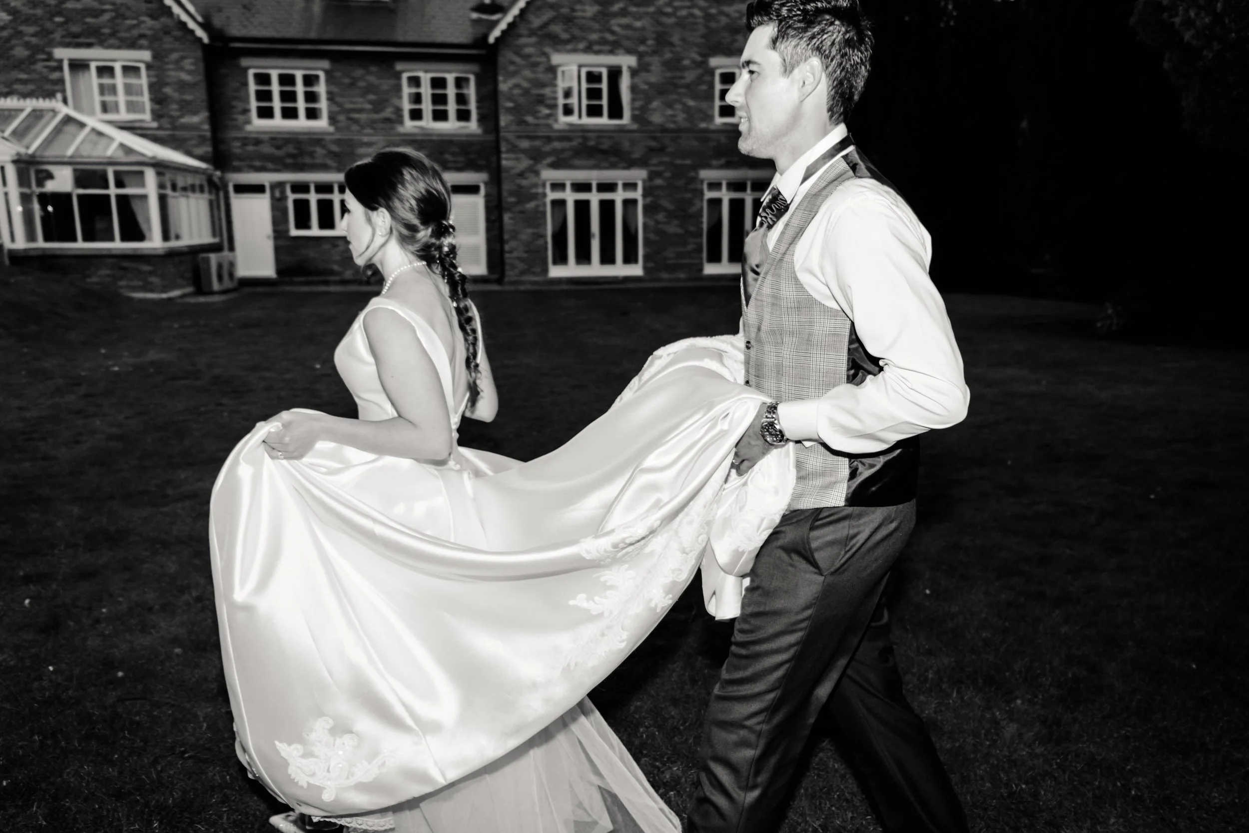 A black and white photo of a bride and groom at night, with the groom carrying the bride in a wedding dress across a grassy area outside a building with multiple windows.