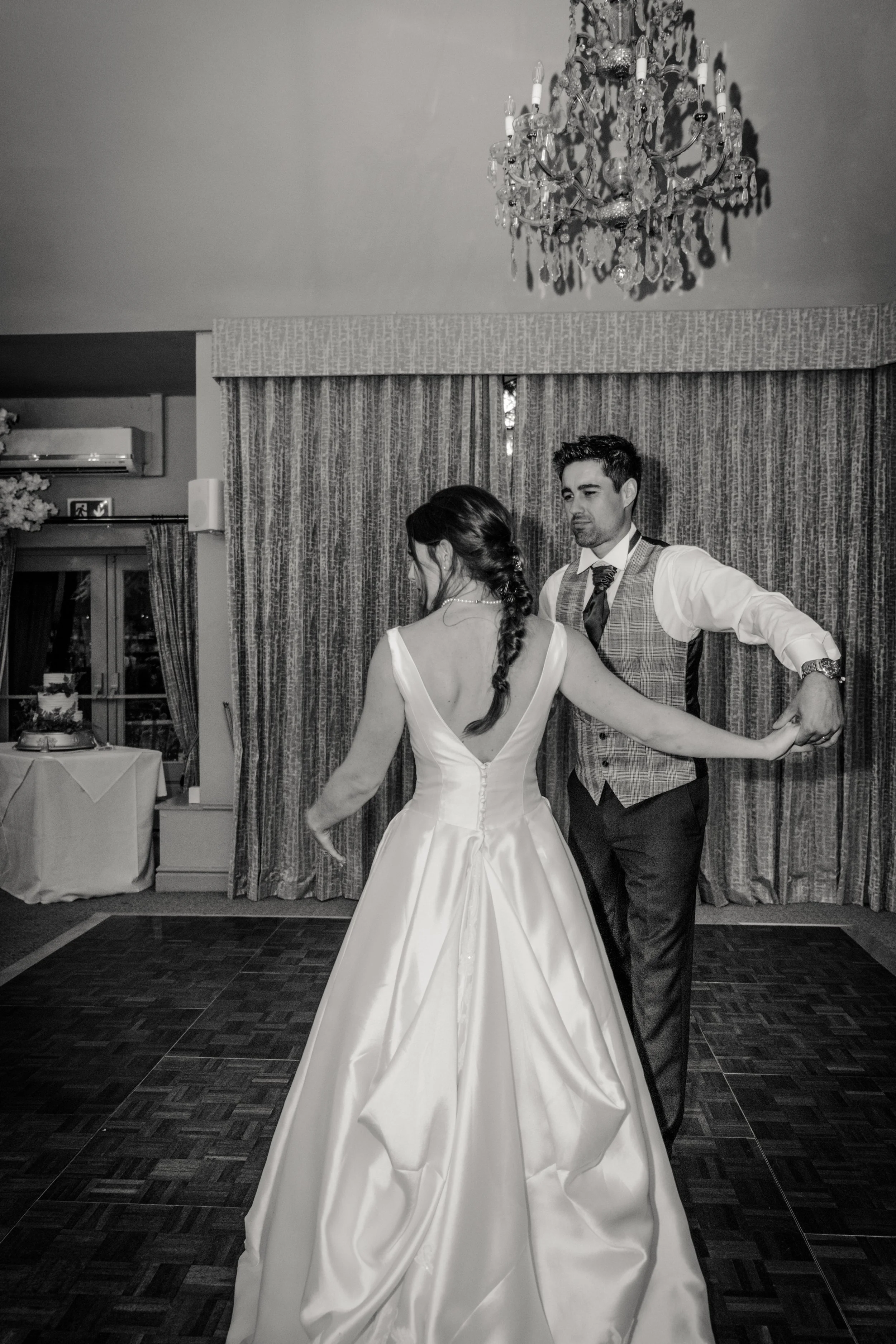 A bride and groom dancing at a wedding reception, with the bride in a white gown and the groom in a vest and tie, in a room with curtains and chandelier.