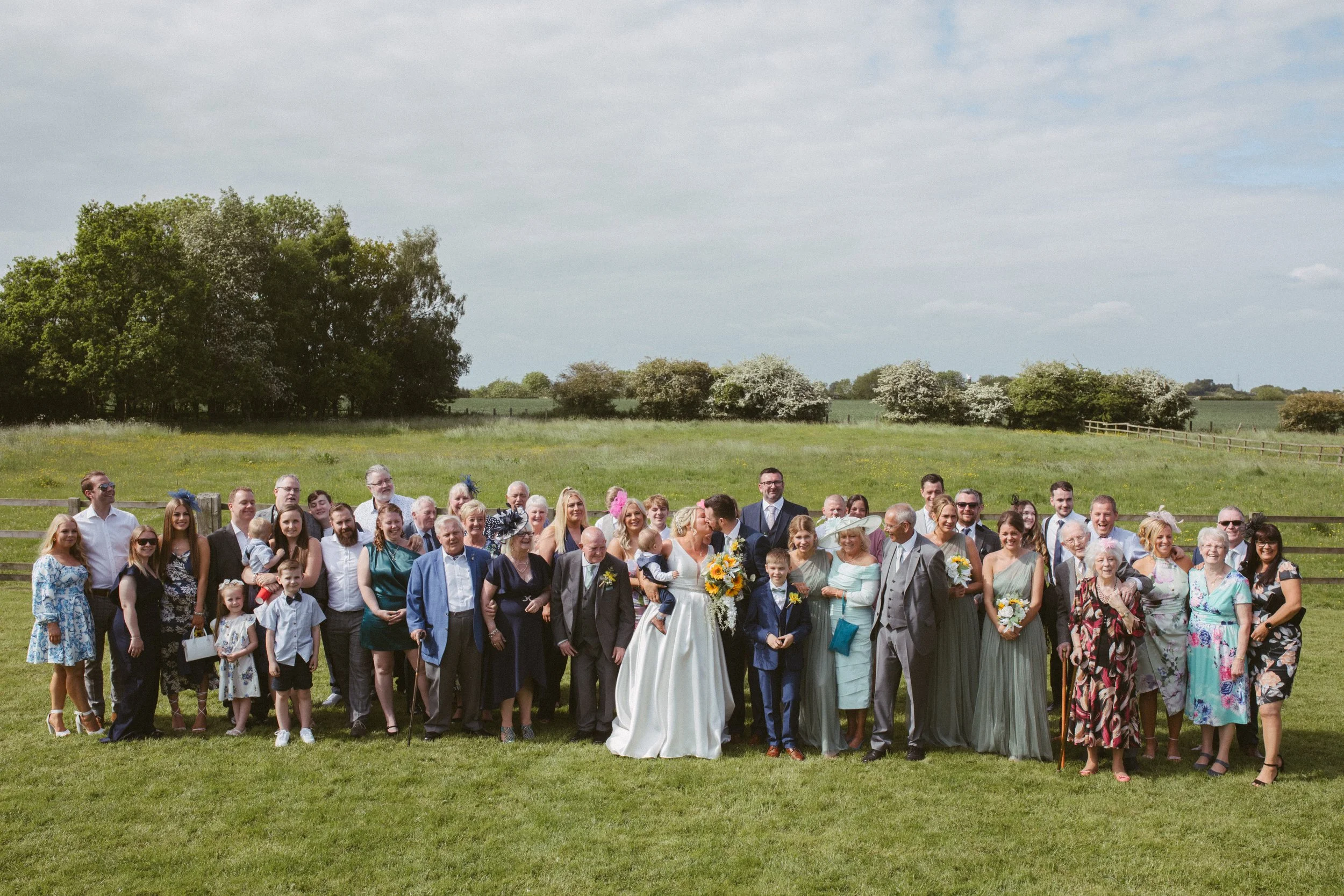 A large group of people gathered outdoors on a grassy field with trees and open sky in the background, likely celebrating a wedding.