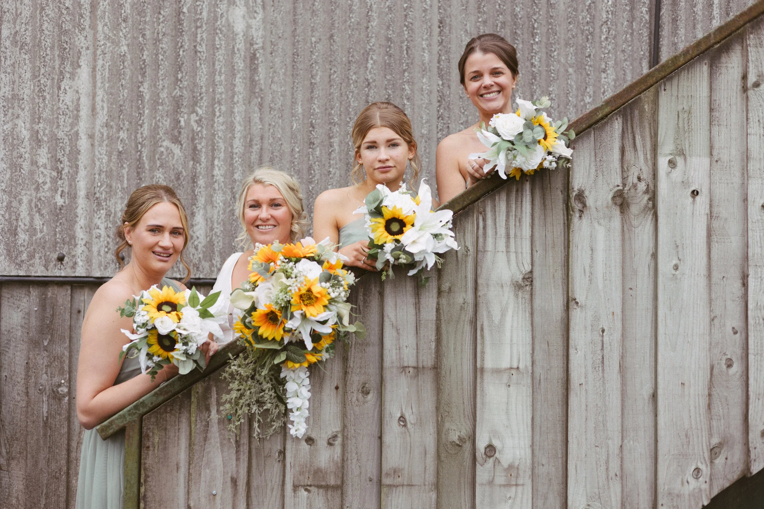 A group of five women in formal dresses holding bouquets of flowers, standing behind a wooden fence, smiling at the camera.