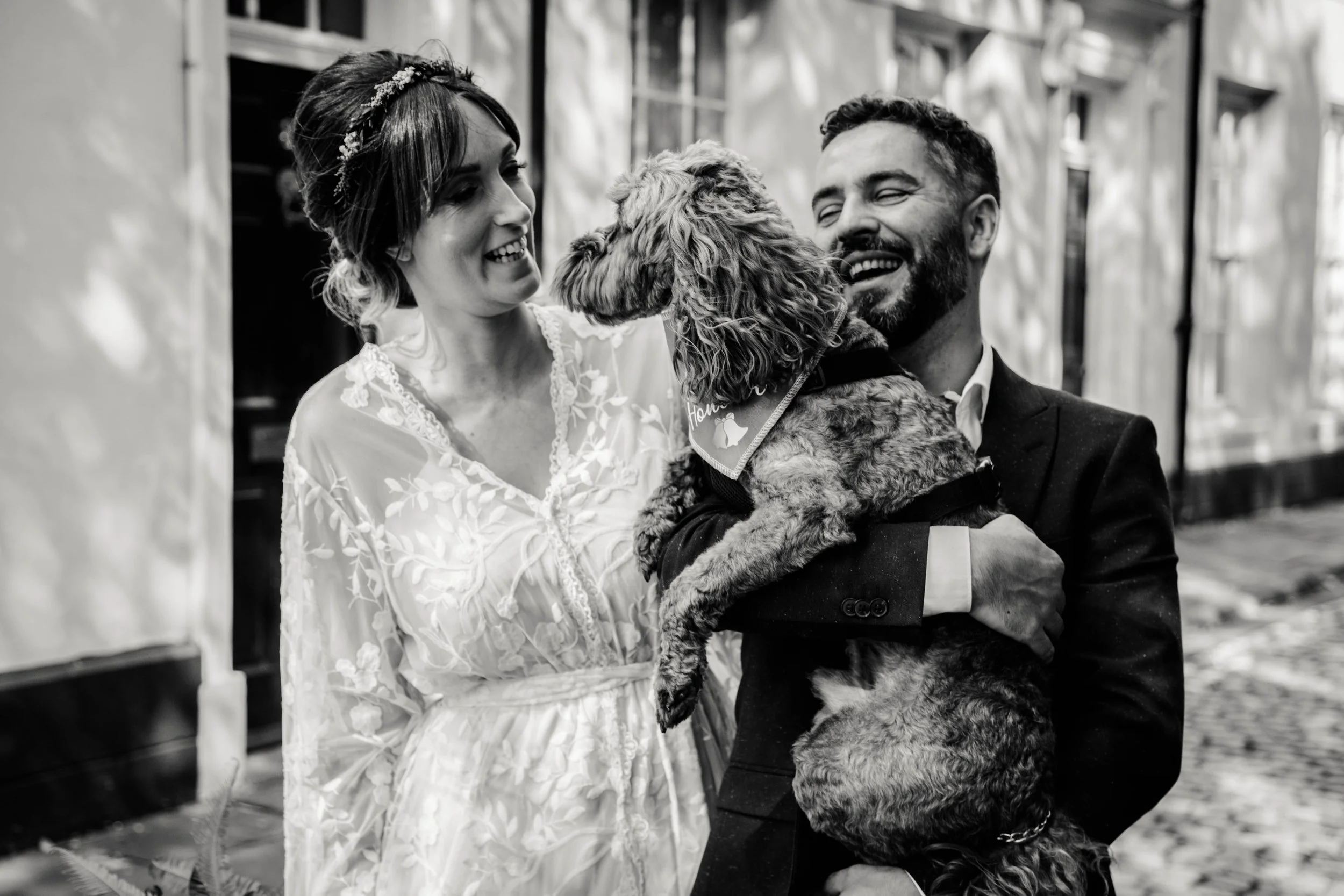 A joyful couple in formal wedding attire with a small dog, celebrating outdoors with an architecture background.