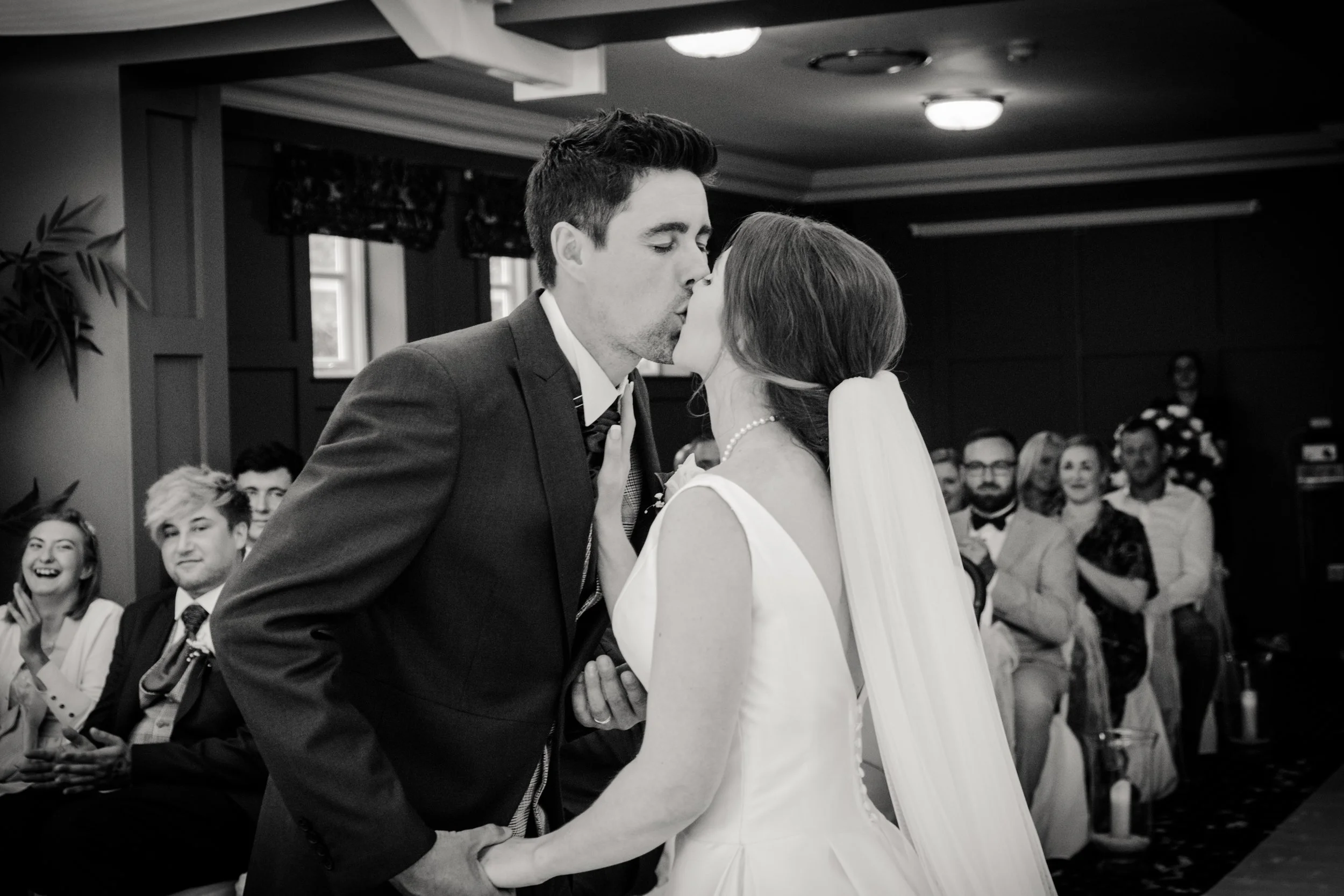 A black-and-white photo of a wedding kiss between a groom in a tuxedo and a bride in a white dress with a pearl necklace, with guests seated in the background, during a wedding ceremony.