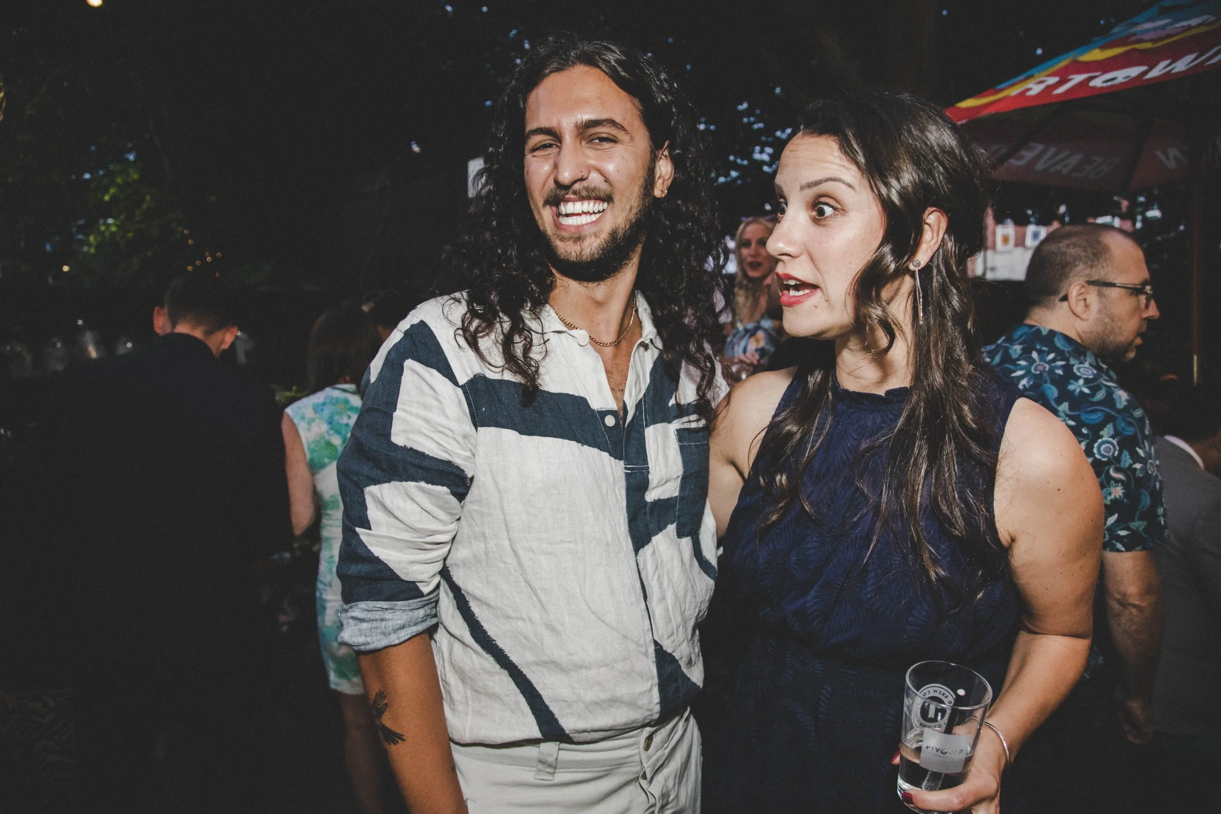 Two young adults, a man with long curly dark hair and a woman with long dark hair, are standing close together at an outdoor event at night, smiling and talking. The man is wearing a striped shirt, and the woman is wearing a dark blue patterned dress