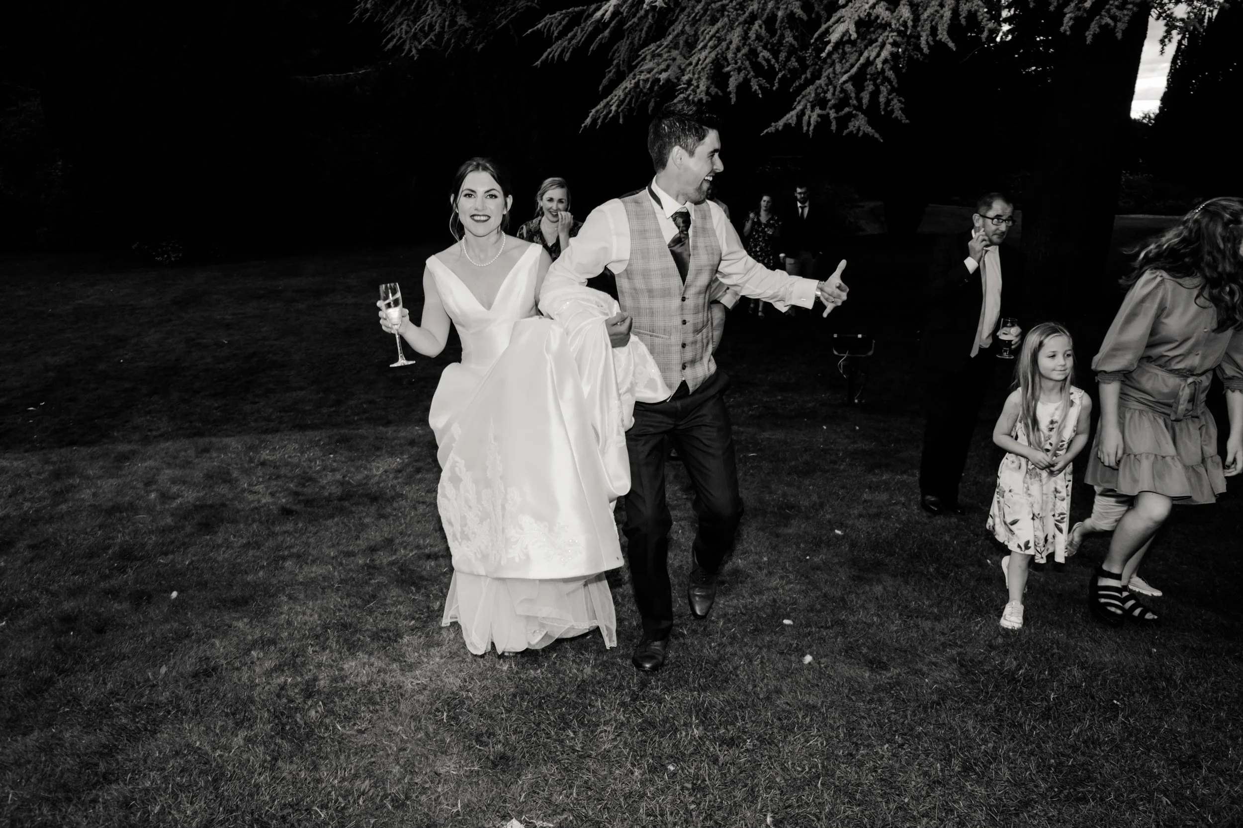 Black and white photo of a wedding celebration outdoors. The bride, in a white wedding gown, is holding a glass and smiling. The groom, in a vest and tie, is giving a thumbs-up. Several guests are visible in the background, including a young girl and