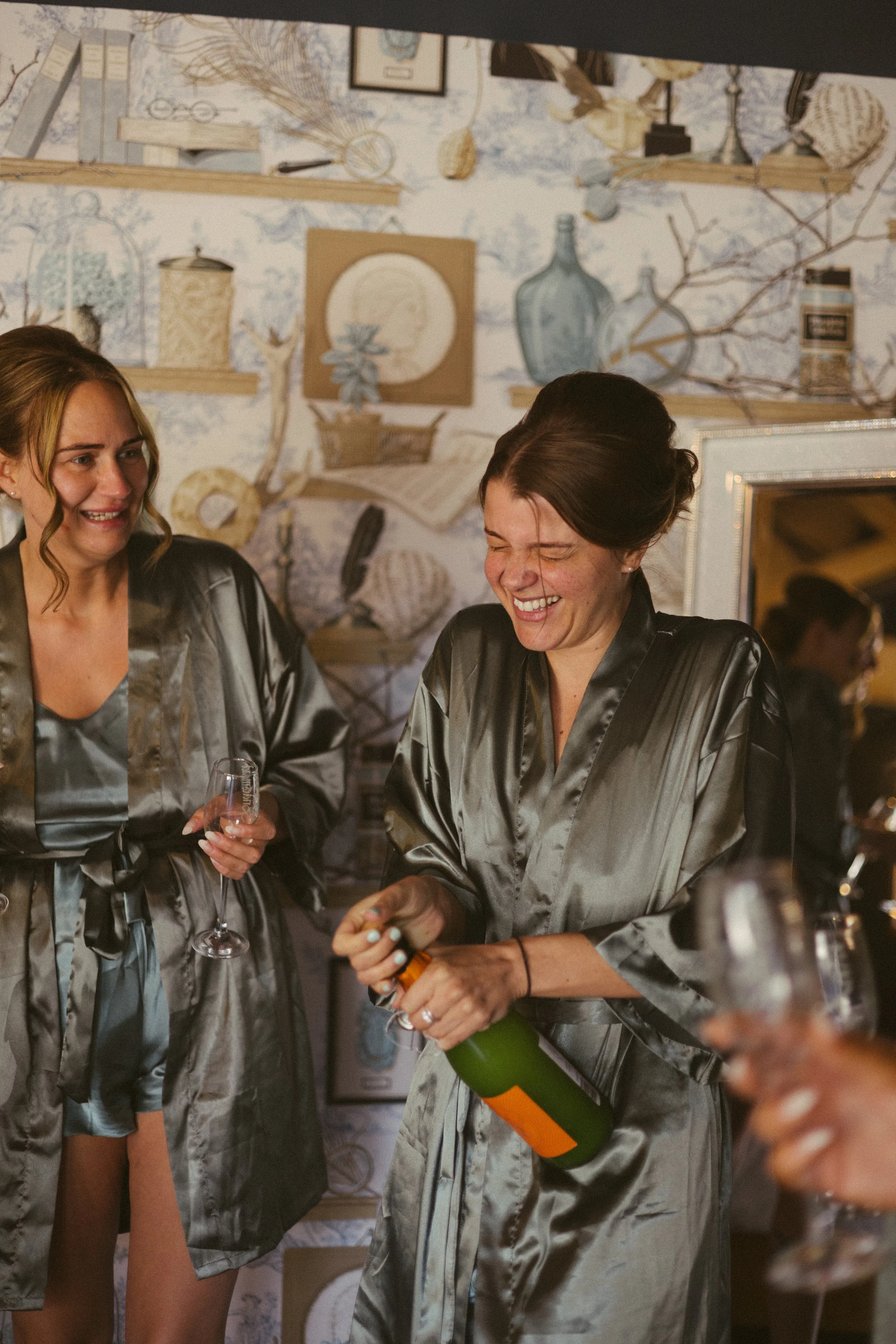 Two women in matching satin robes celebrating, one holding a glass of champagne and the other opening a bottle of champagne, inside a room with decorative wall art.