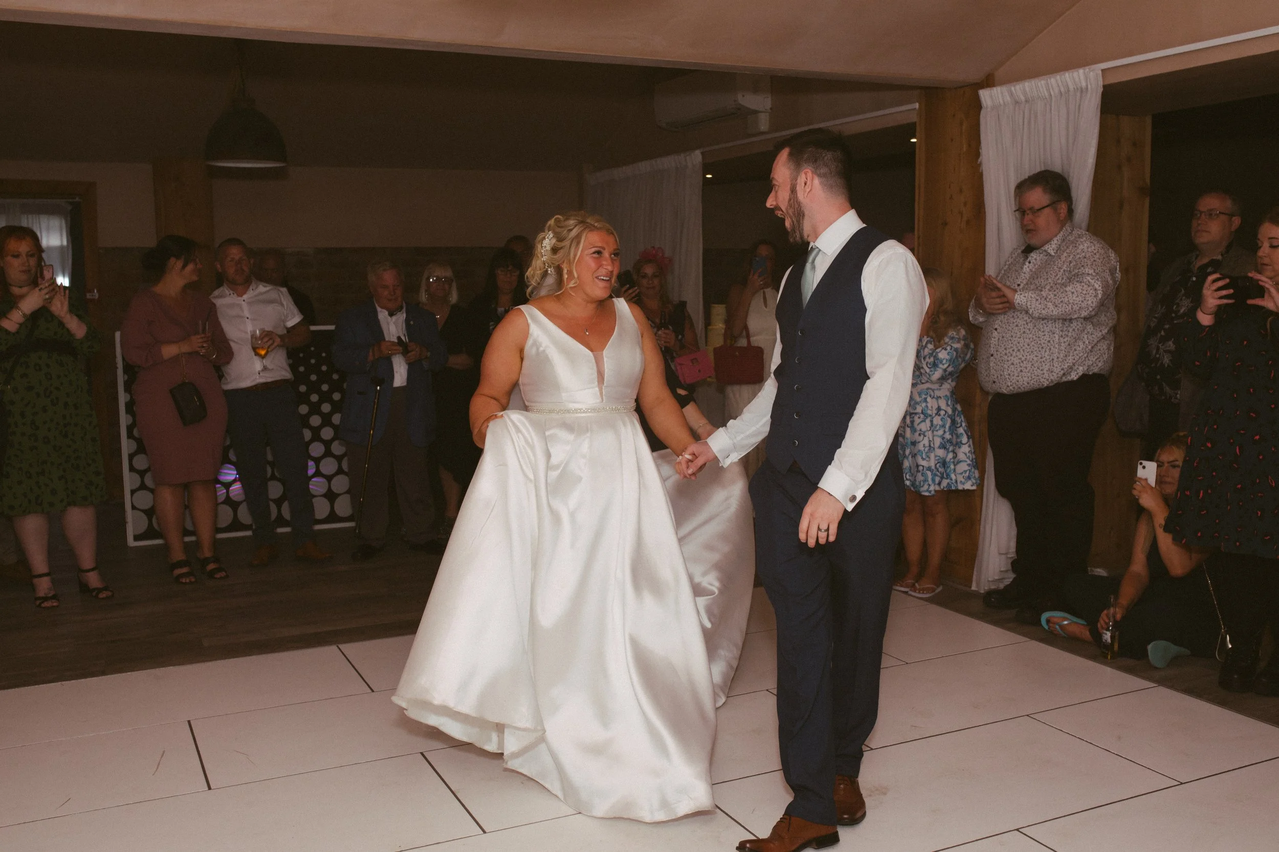 A bride and groom dancing at their wedding reception, surrounded by guests watching and taking photos.
