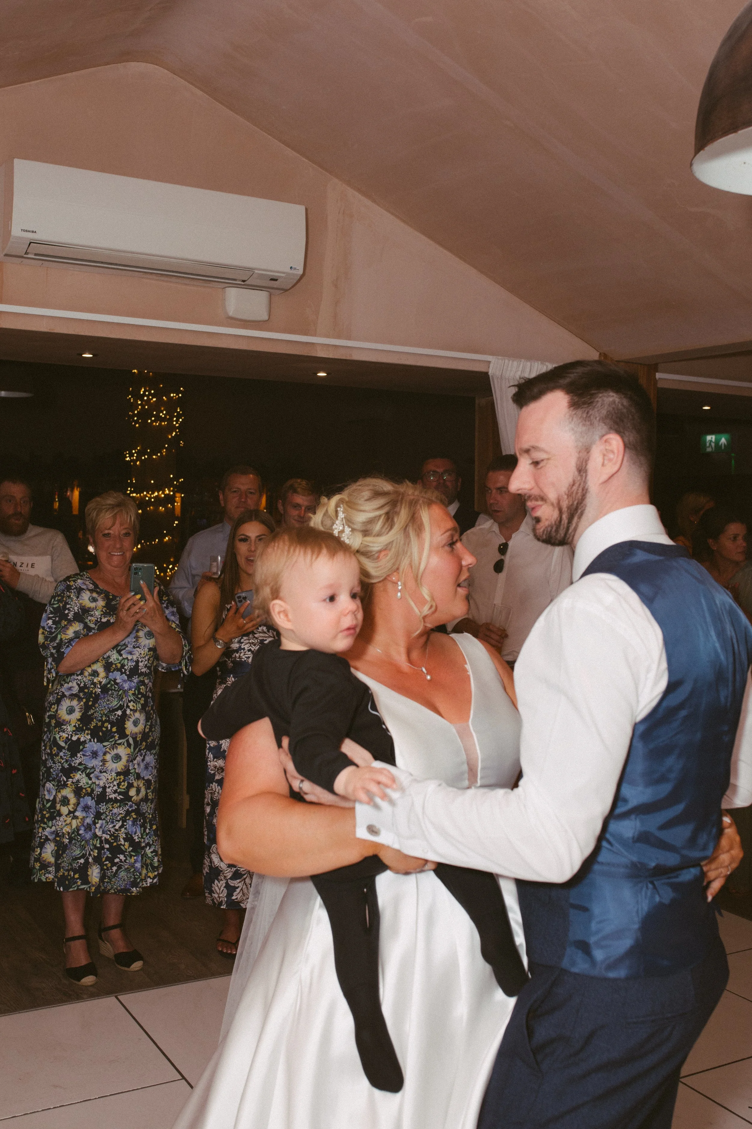 A couple dancing closely at a celebration, with an onlookers group in the background, indoors with warm lighting and a Christmas tree.