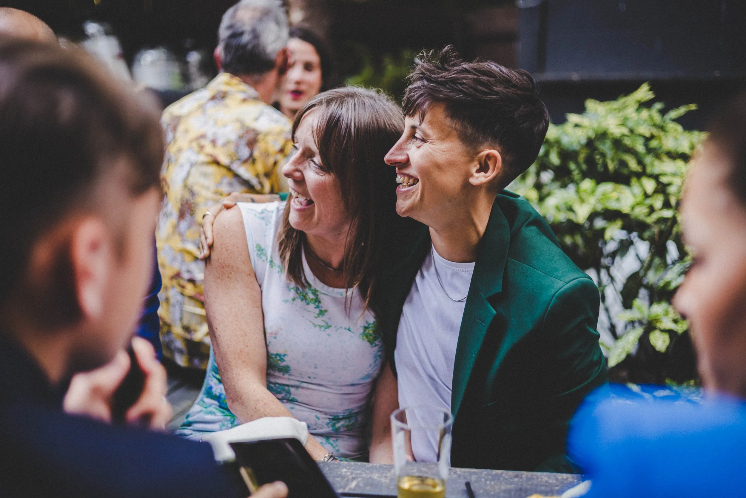 People smiling and socializing at a gathering, with a woman and a young man in focus, surrounded by others in a lively outdoor setting with greenery.